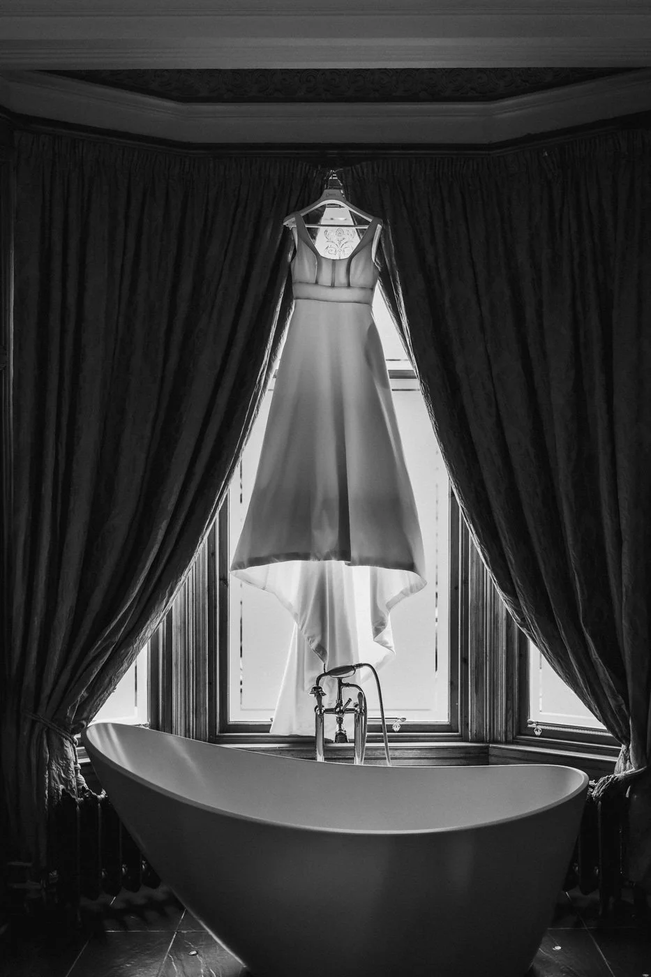Wedding dress hanging in front of a window with a clawfoot bathtub below.