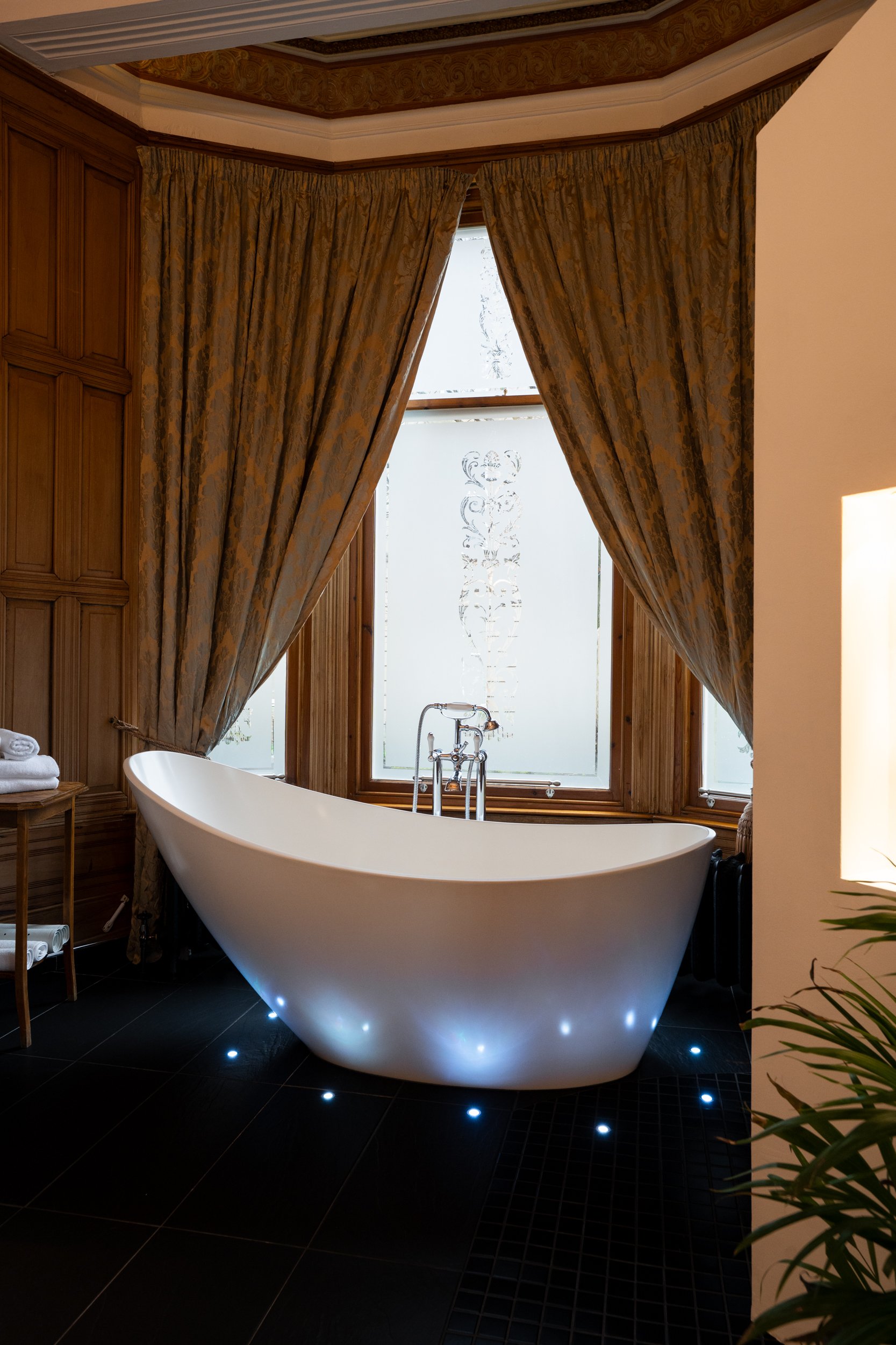A modern bathroom with a white free-standing bathtub, illuminated by small embedded floor lights, situated in front of frosted windows with decorative patterns, framed by gold-patterned curtains, and wooden wall panels.