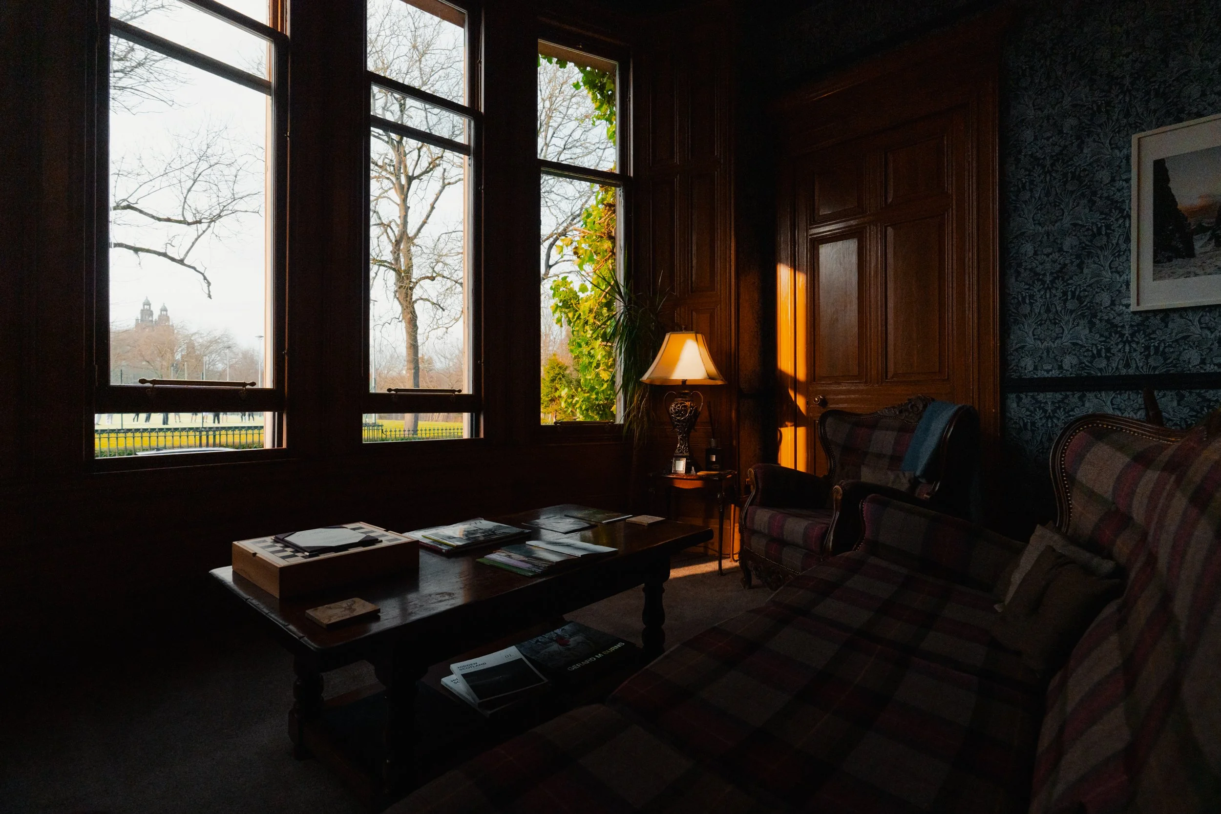 A cozy living room with dark wood paneling, a window with a view of trees outside, a table with books, a plaid sofa and armchair, a lamp on a small table, and framed artwork on the wall.