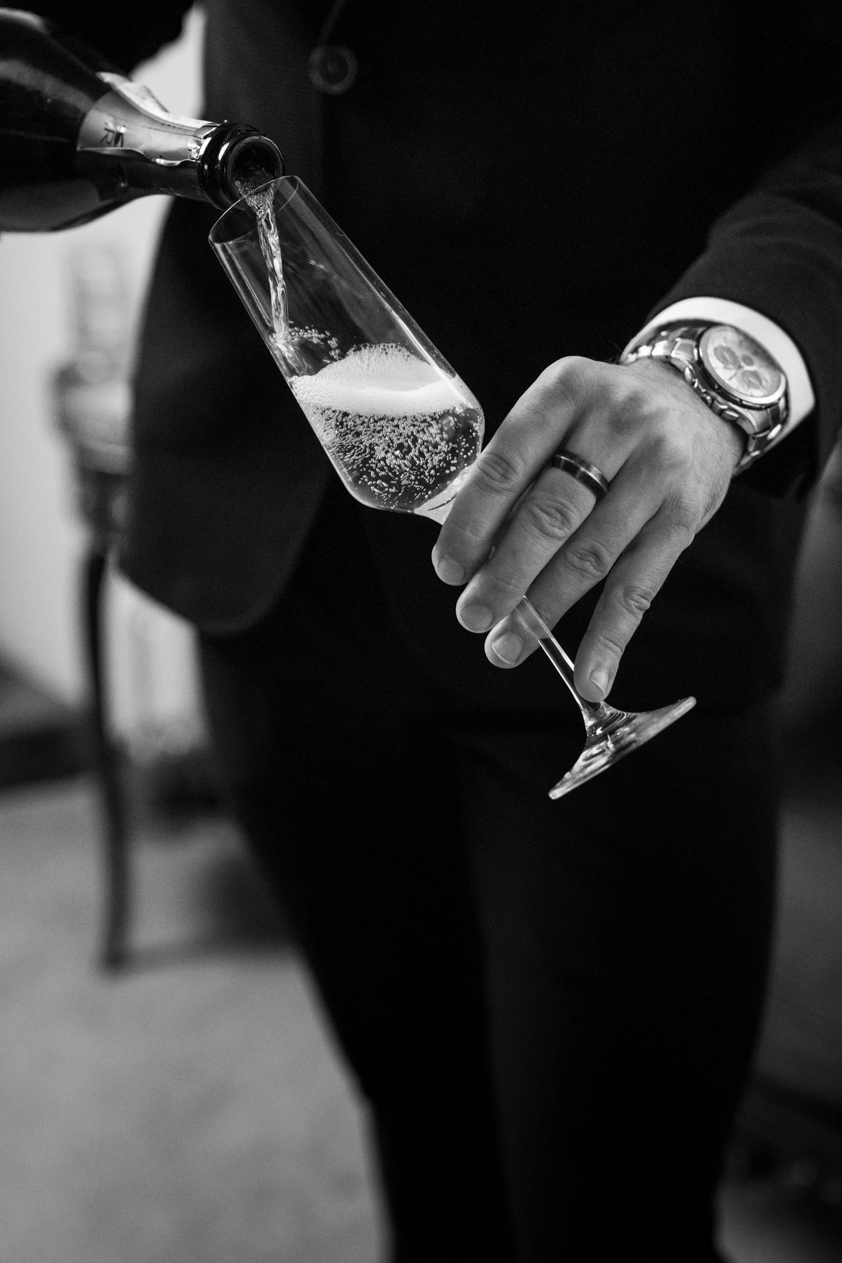 A person in formal attire pours champagne into a flute. The person is wearing a watch and a ring.