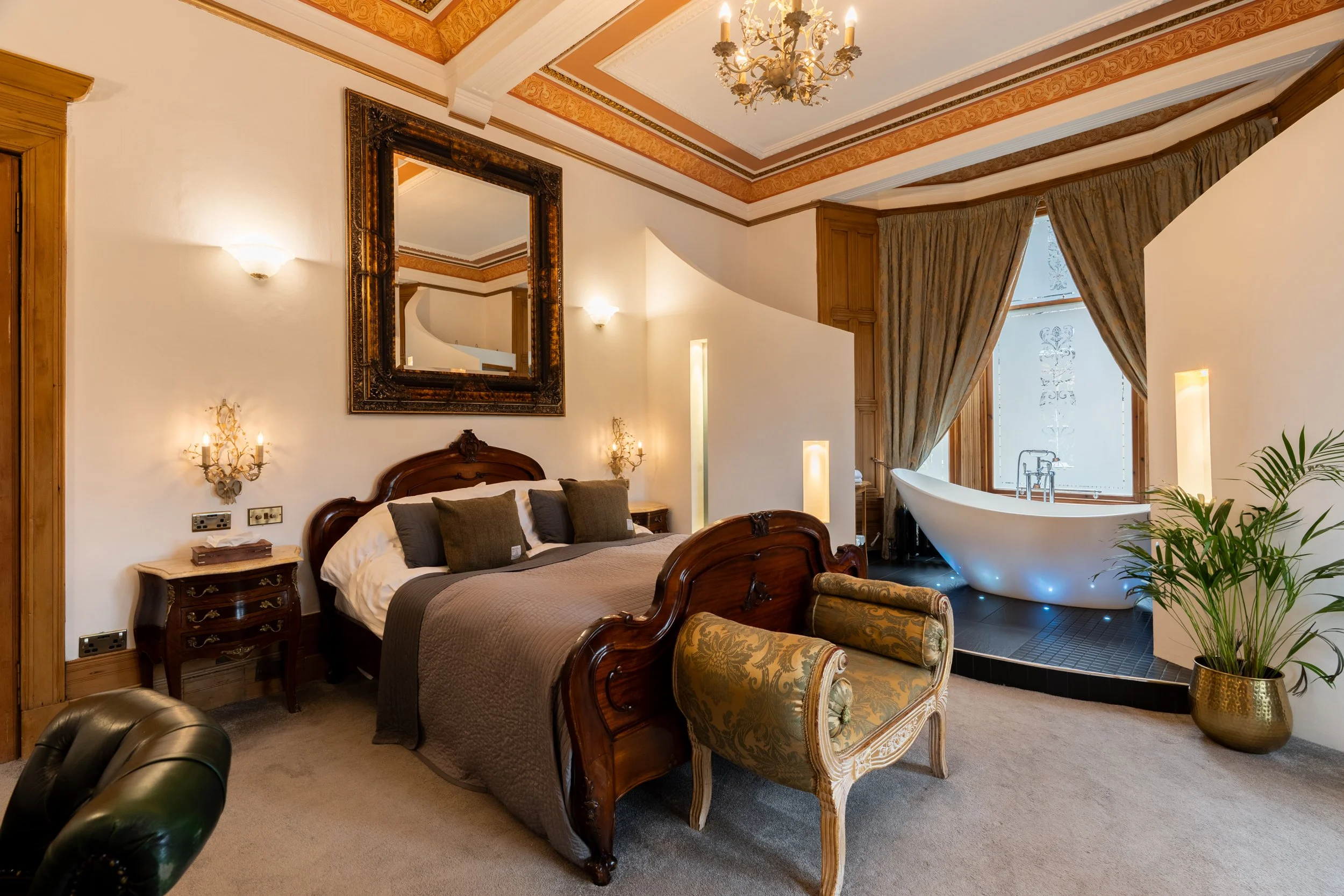 Luxury bedroom with a wooden bed, green cushions, a vintage armchair, and a freestanding bathtub near large windows with curtains.