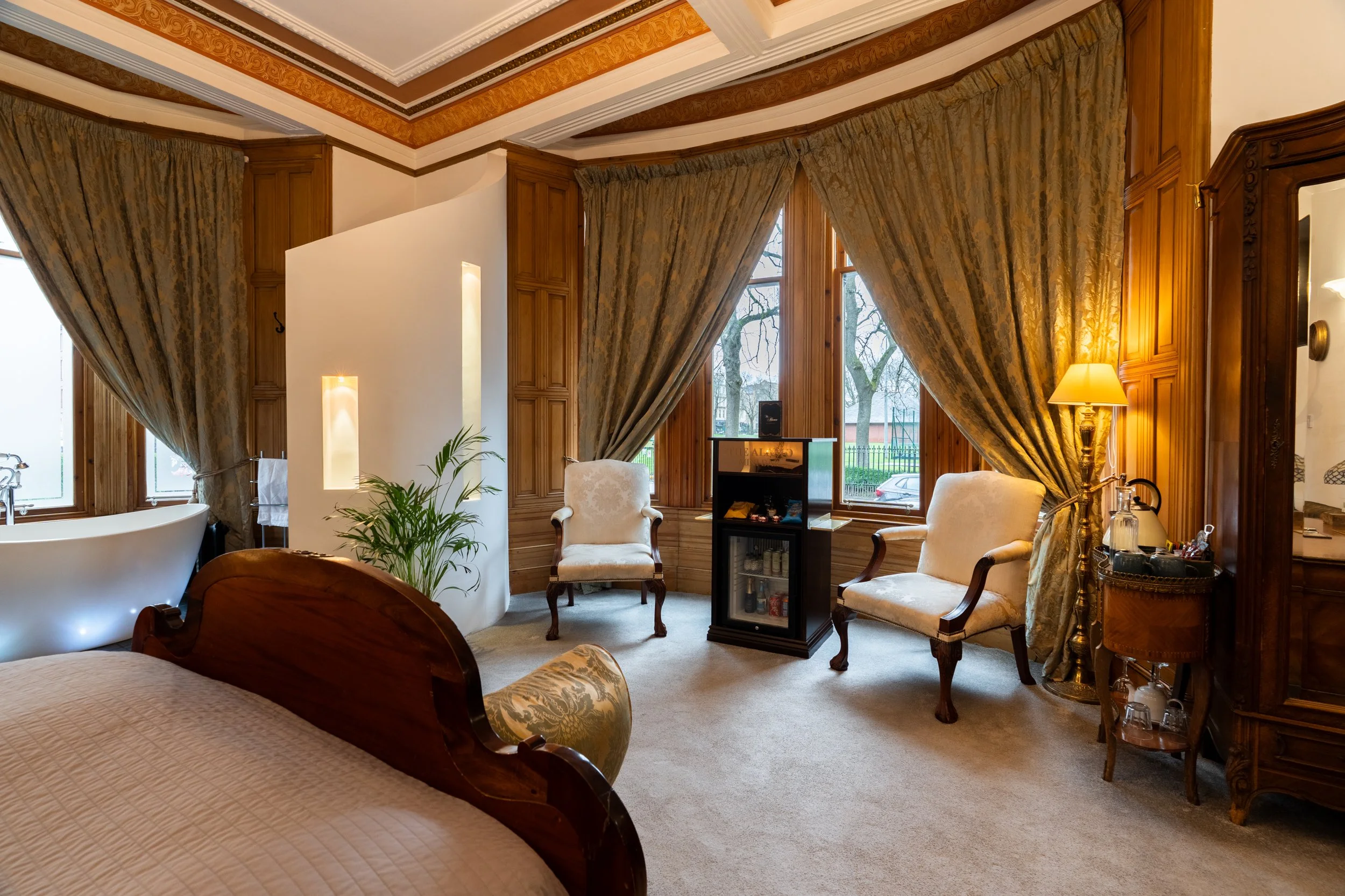 A luxurious bedroom with large windows, gold-patterned curtains, antique furniture including two armchairs, and a freestanding bathtub near the window.