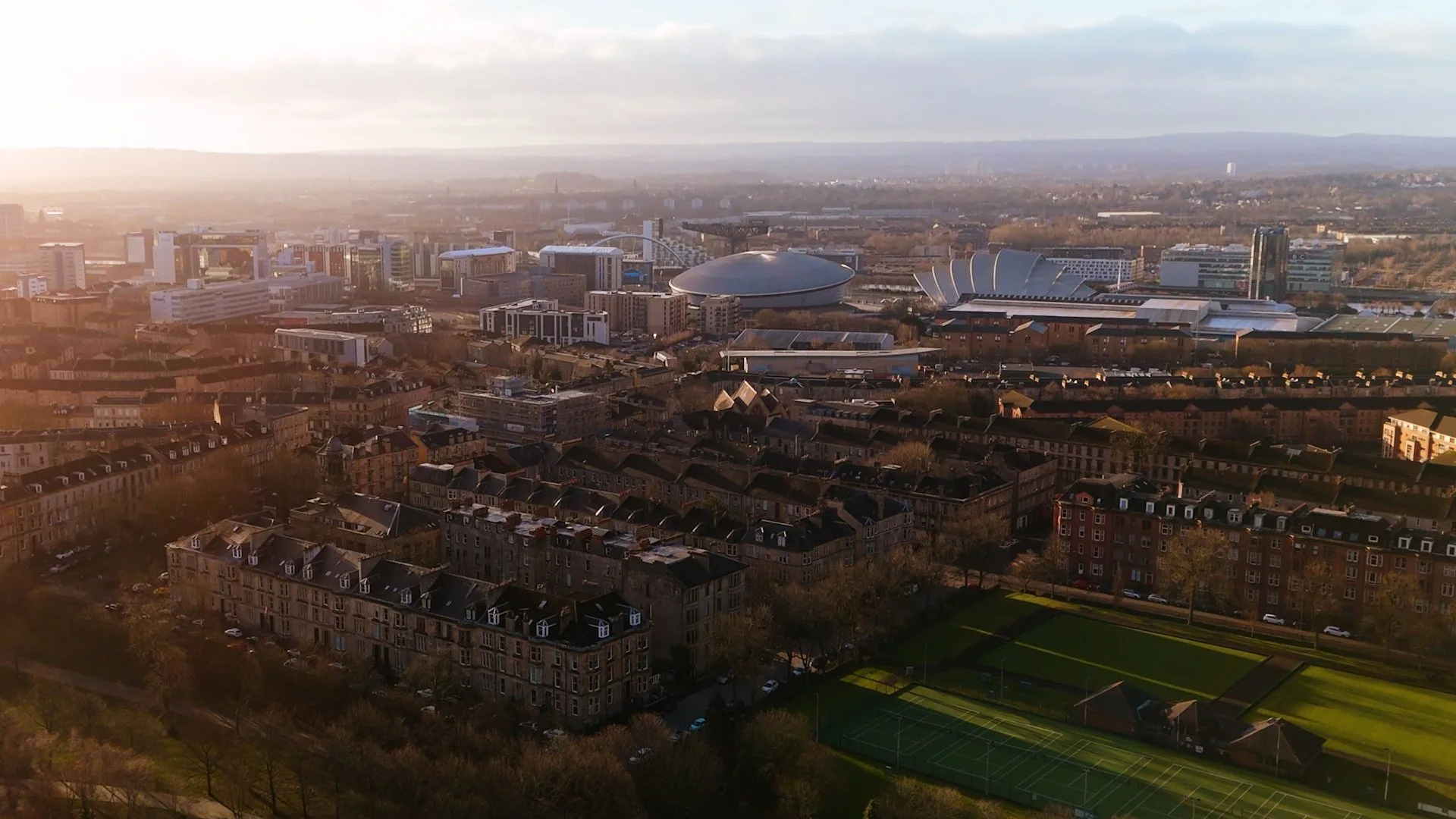 Aerial view of a cityscape at sunset showing a mix of residential, commercial, and recreational areas with a large sports field in the foreground.