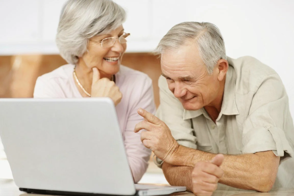 An elderly couple smiling and looking at a laptop screen together, sitting at a table.