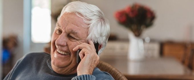 Older man smiling while talking on a cell phone indoor with a blurred background.