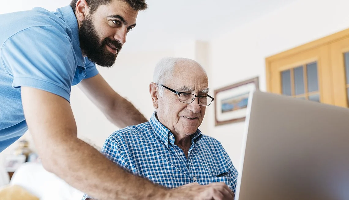 A young man helping an elderly man use a laptop in a home setting.