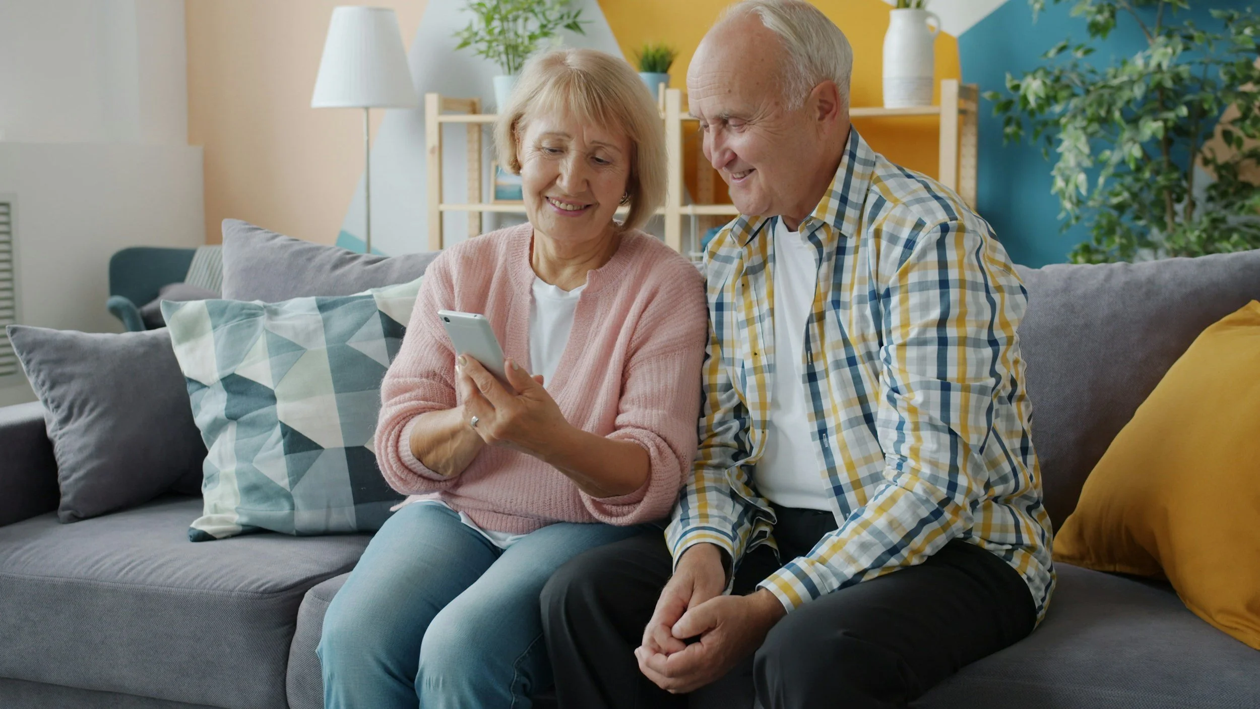 An elderly couple sitting on a gray couch, looking at a smartphone together, smiling in a cozy living room.