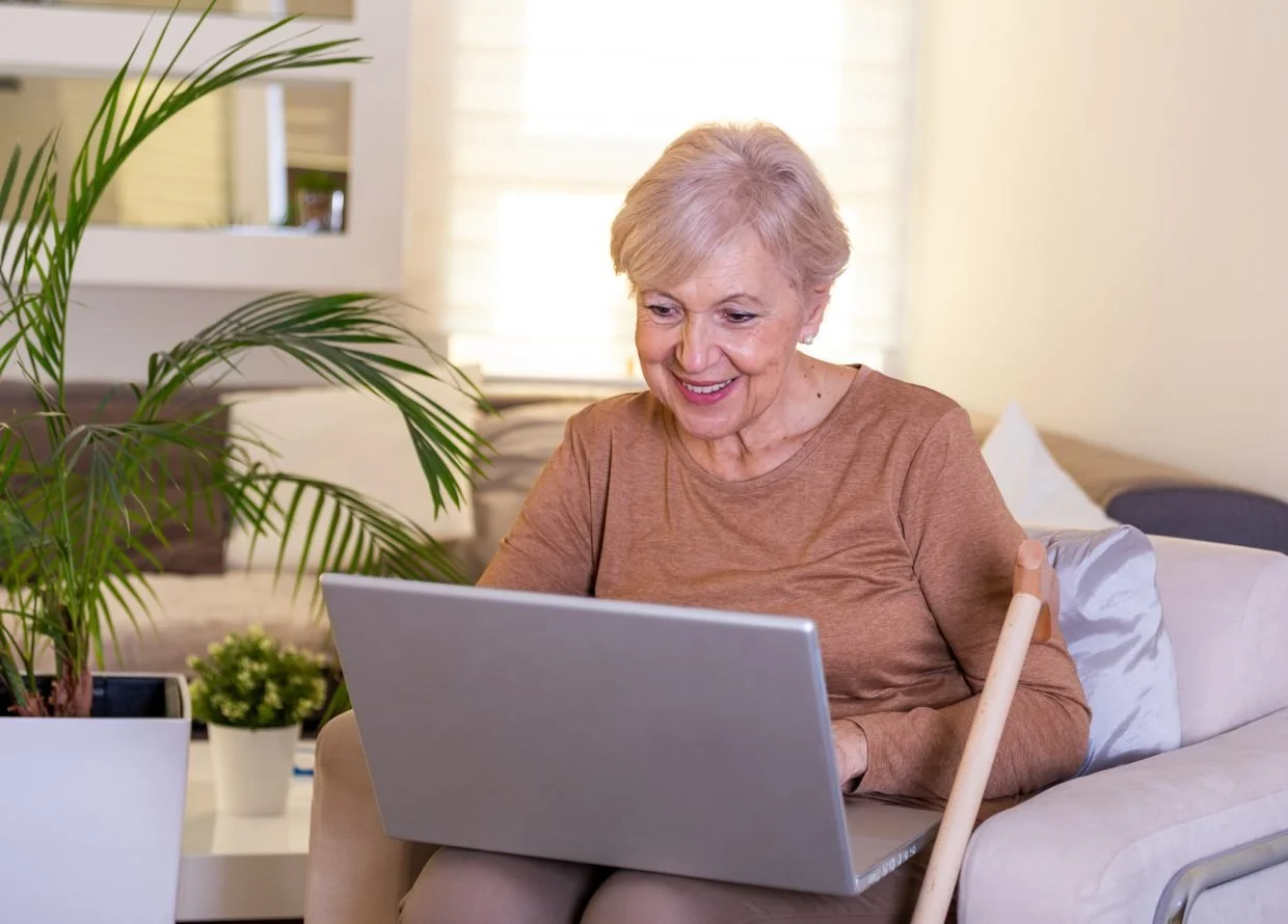 An elderly woman with short gray hair sitting on a sofa, working on a laptop, smiling, with a cane resting next to her, in a bright living room with plants and decorative pillows.