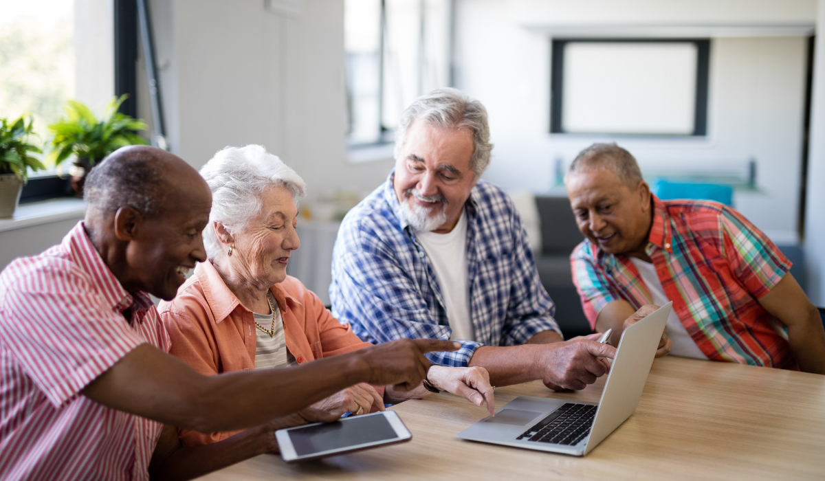 Four elderly people sitting at a table, smiling and looking at a laptop, with one showing something on their phone, in a bright room with plants and windows.