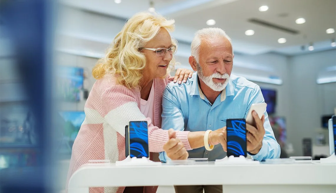 An elderly woman and man in a store, looking at a smartphone together, with the woman showing interest and the man holding the phone, surrounded by display devices.