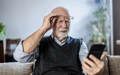 An elderly man with glasses, wearing a gray sweater vest over a light blue shirt, looking confused at his smartphone while sitting on a couch in a living room.