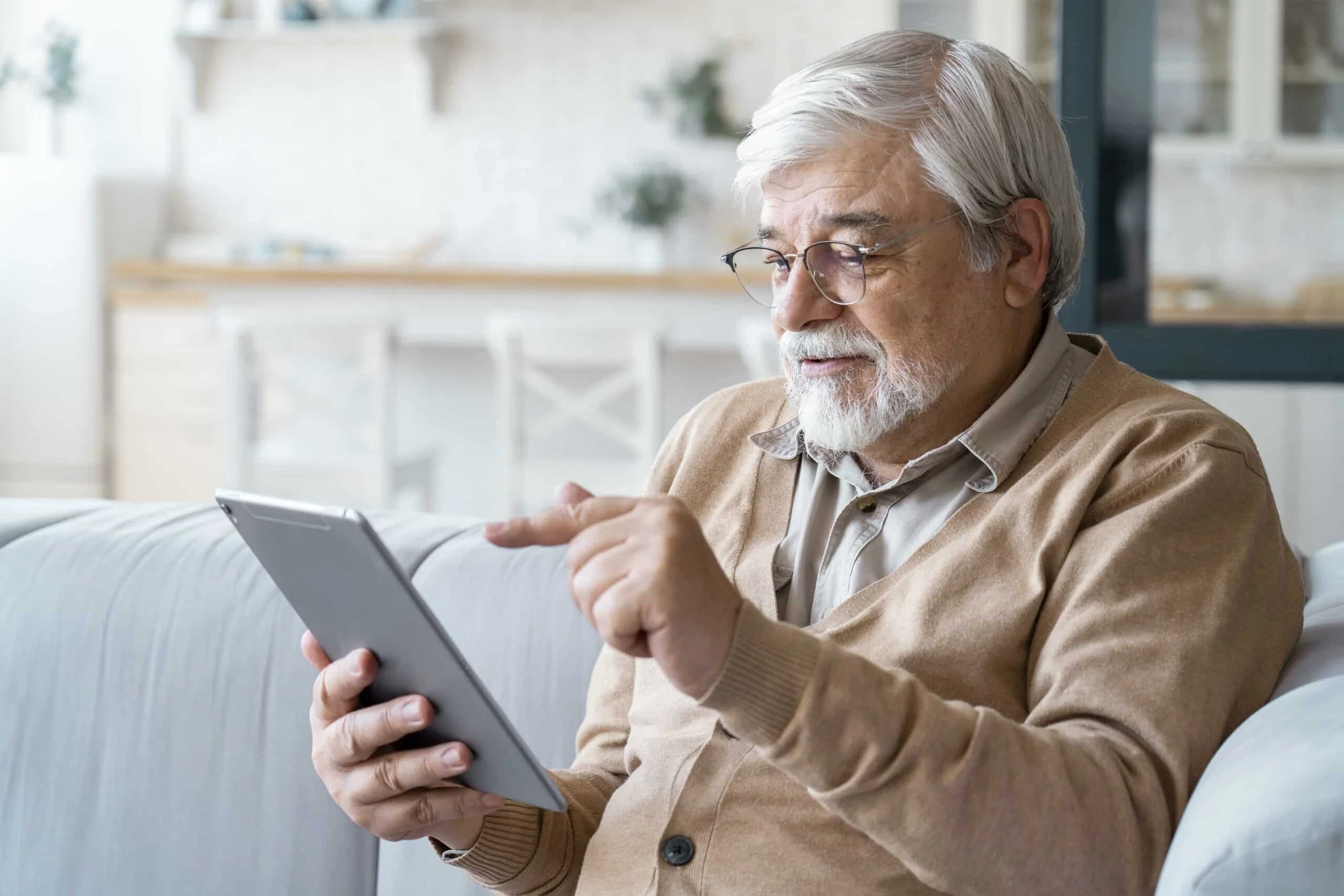 An elderly man with gray hair and a beard, wearing glasses, a beige cardigan, and a light shirt, sitting on a sofa and using a tablet in a bright, modern living room.