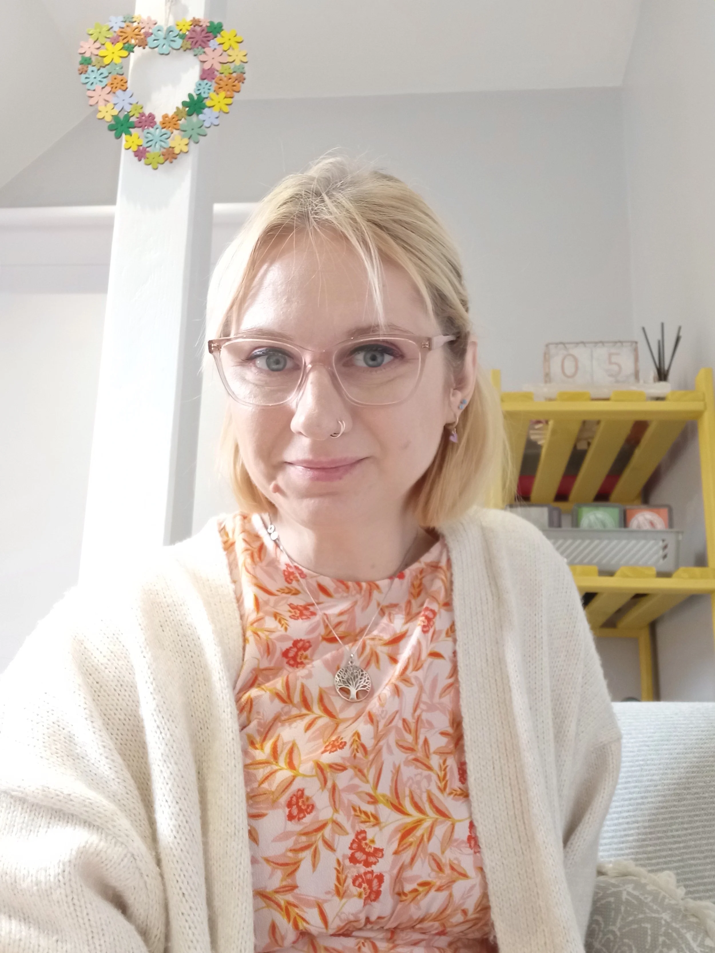 A woman with blonde hair, glasses, and a nose piercing is smiling for a selfie. She is wearing a floral dress, a cream cardigan, and a silver necklace with a tree pendant. Behind her, a yellow shelf holds decorative items, and a heart-shaped floral wreath hangs above a white support beam.