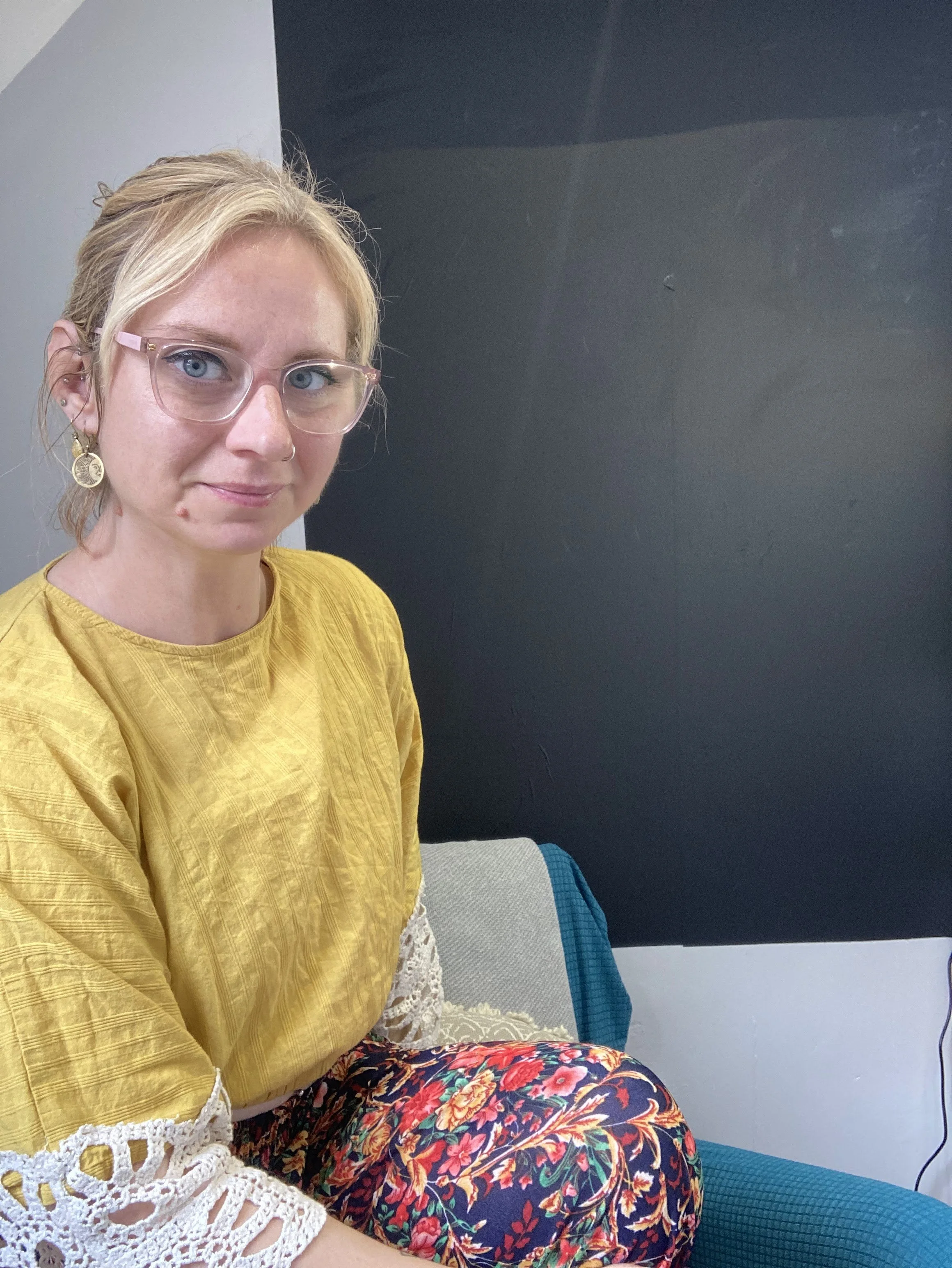 A woman with blonde hair and glasses sitting on a chair, wearing a yellow top and floral pants, with earrings and a nose ring, against a background of a dark and light wall.