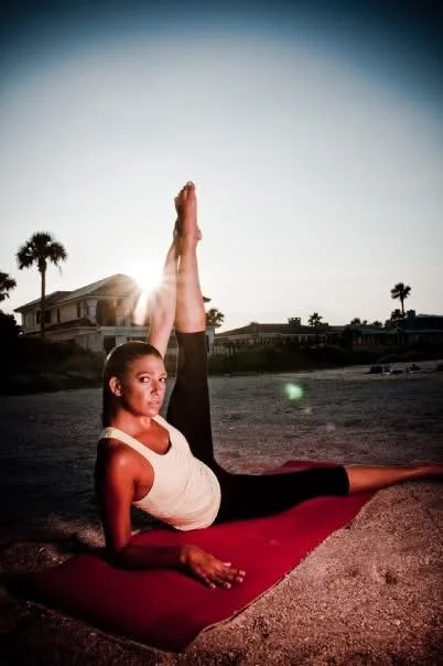 A woman practicing yoga outdoors on a mat during sunset, performing an extended side stretch pose, with palm trees and houses in the background.