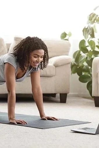 A woman exercising at home on a yoga mat while looking at a laptop.