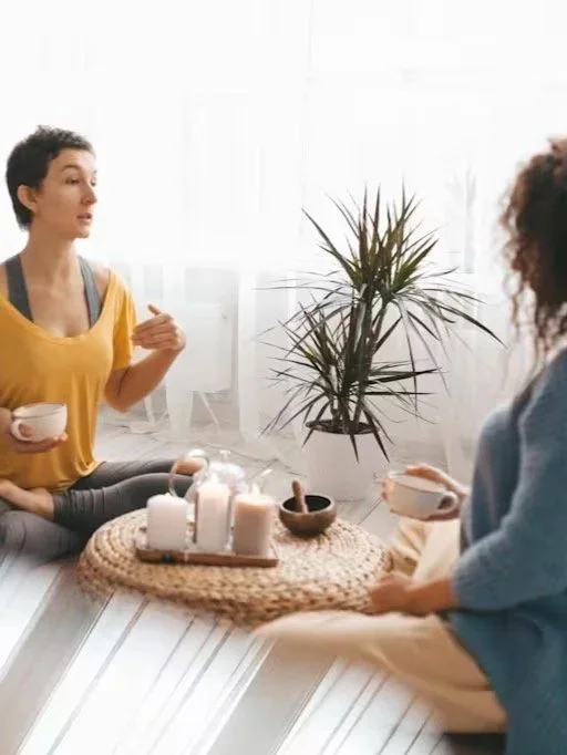 Two women sitting on a white windowsill having a conversation, holding tea cups, with a potted plant and candles on a woven mat between them.