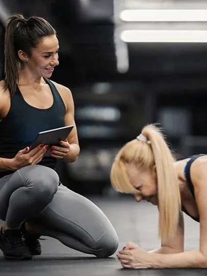 A woman with long dark hair in a ponytail, wearing a black tank top and gray leggings, kneeling and holding a notepad while smiling. A blonde woman with long hair in a ponytail, wearing a black sports bra, is doing push-ups on the gym floor.