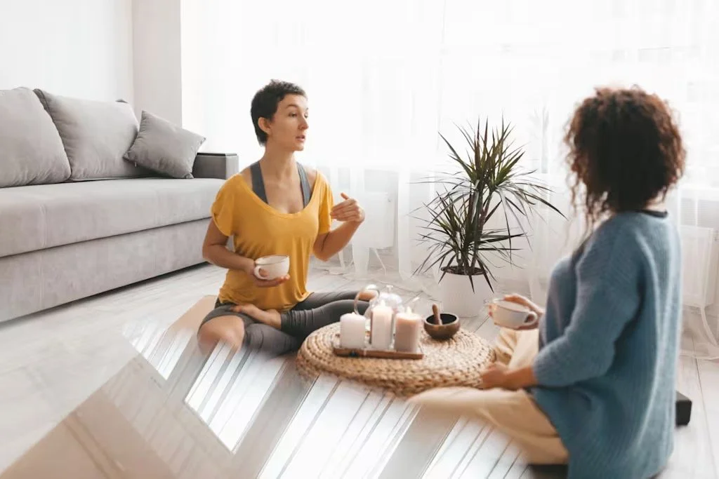 Two women sitting on a beige rug having a conversation. They are holding cups and sitting near a coffee table with candles and bowls. A sofa with pillows and a potted plant are in the background.