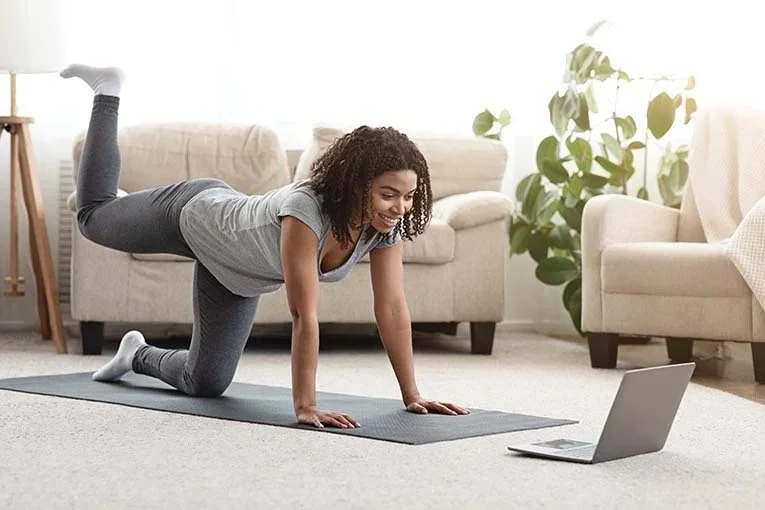 Woman doing a yoga pose on a mat indoors while participating in a virtual workout session via laptop.