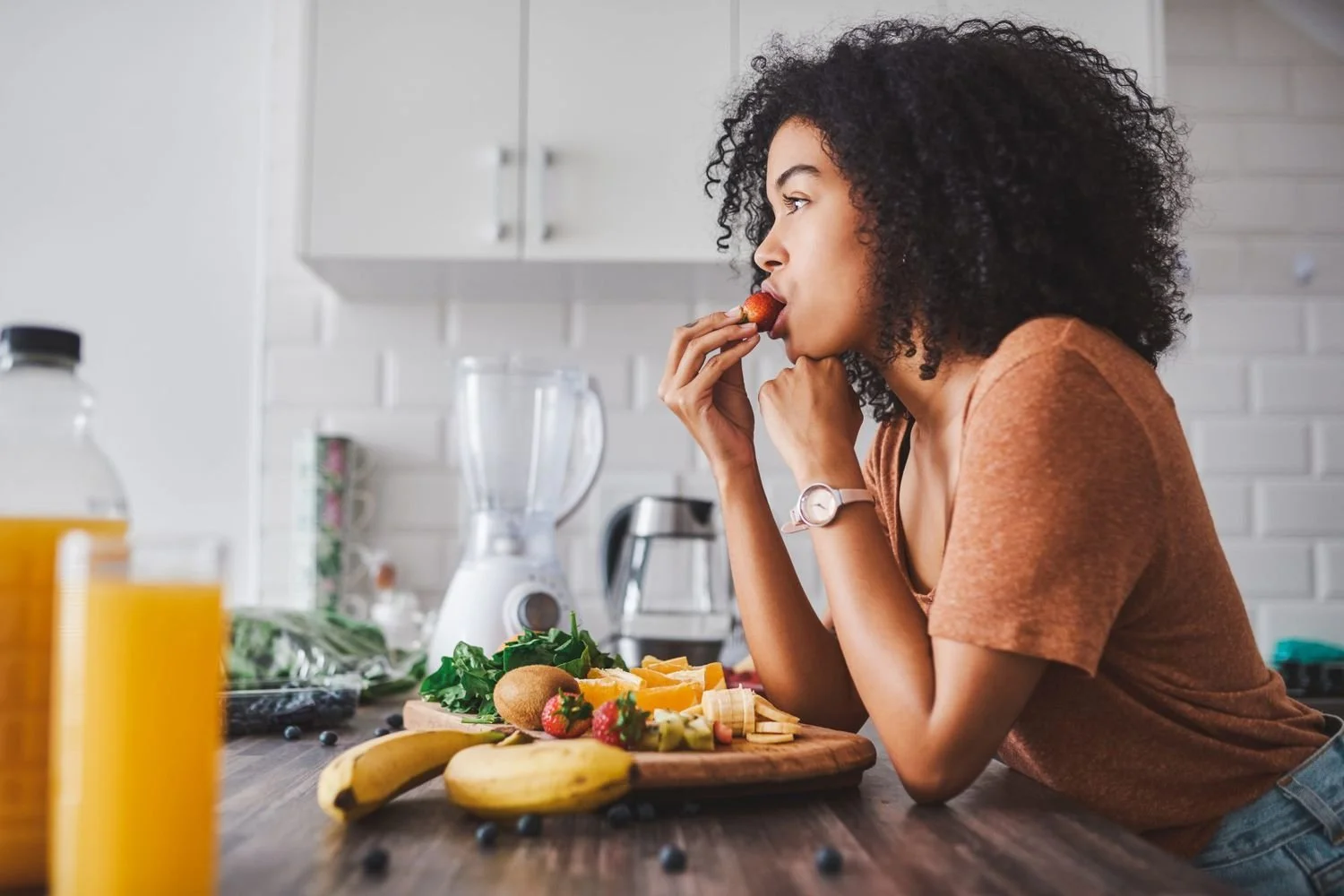 A woman with curly hair eating a strawberry in a modern kitchen with a cutting board of fruit, a blender, and a bottle of orange juice on the counter.
