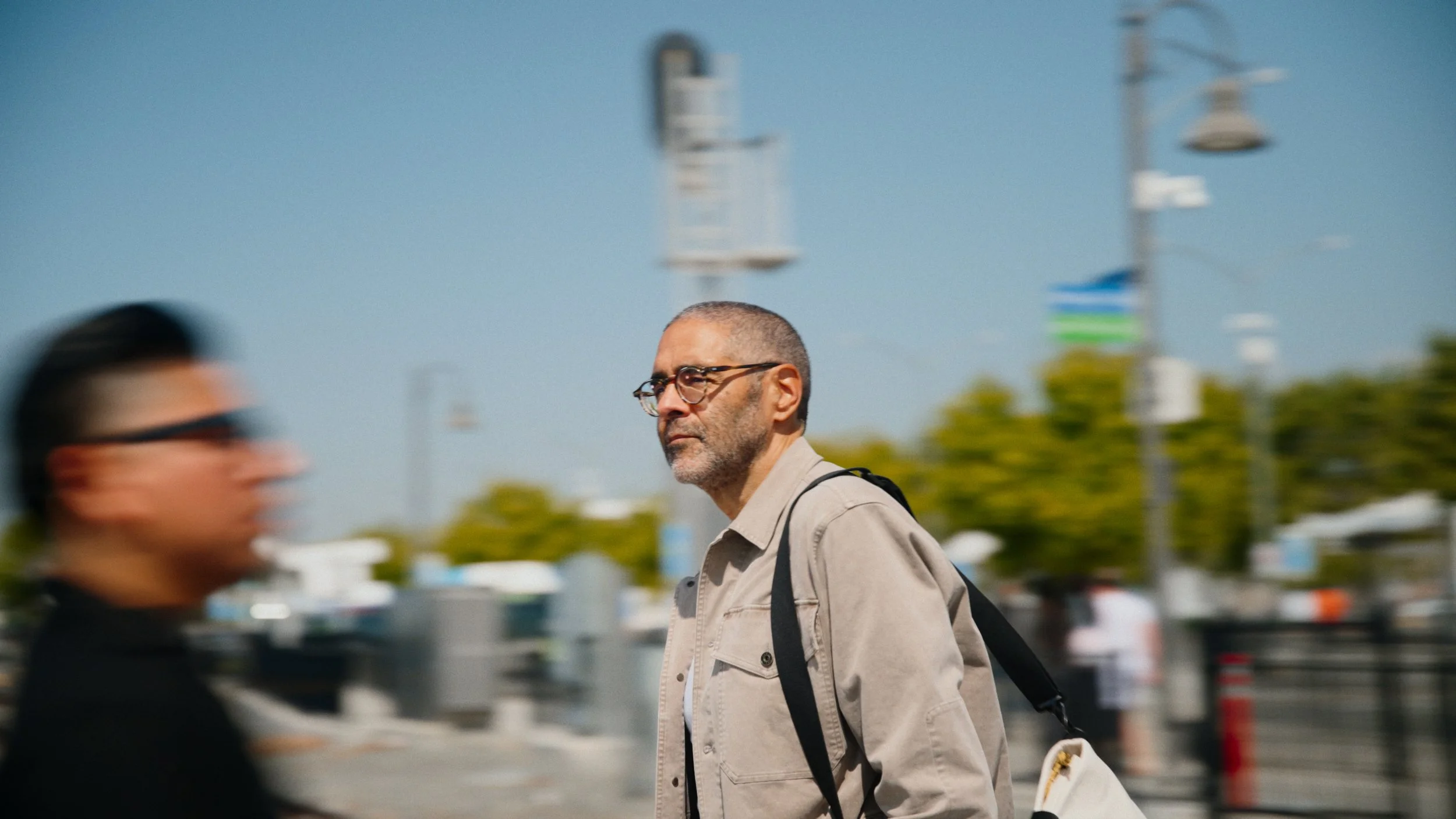 A man with glasses and a beige jacket walking outdoors, with another person blurred in motion in the foreground, on a street or promenade with trees and light poles visible in the background.