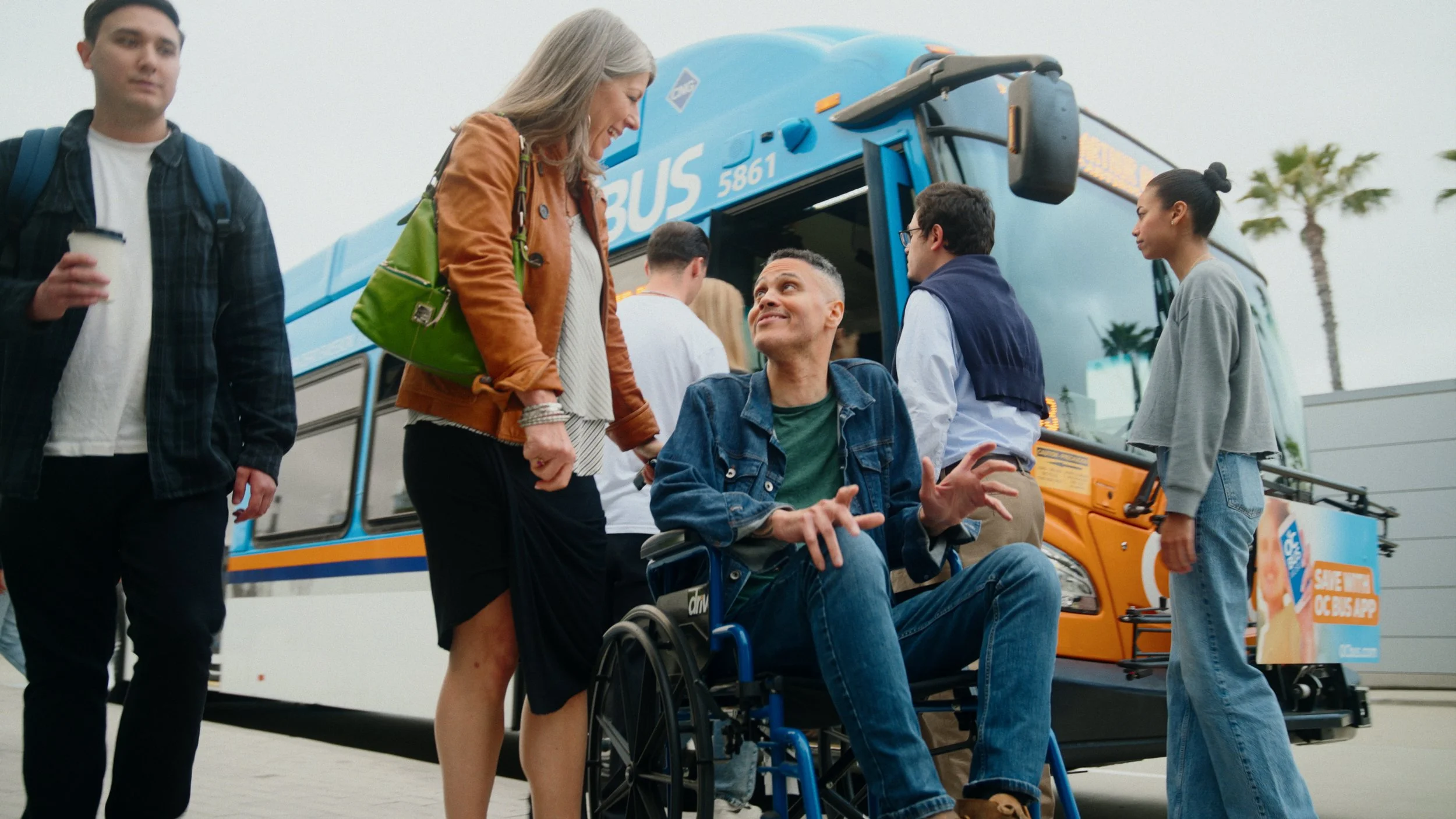A diverse group of people, including a man in a wheelchair, are getting off a blue bus with palm trees in the background.