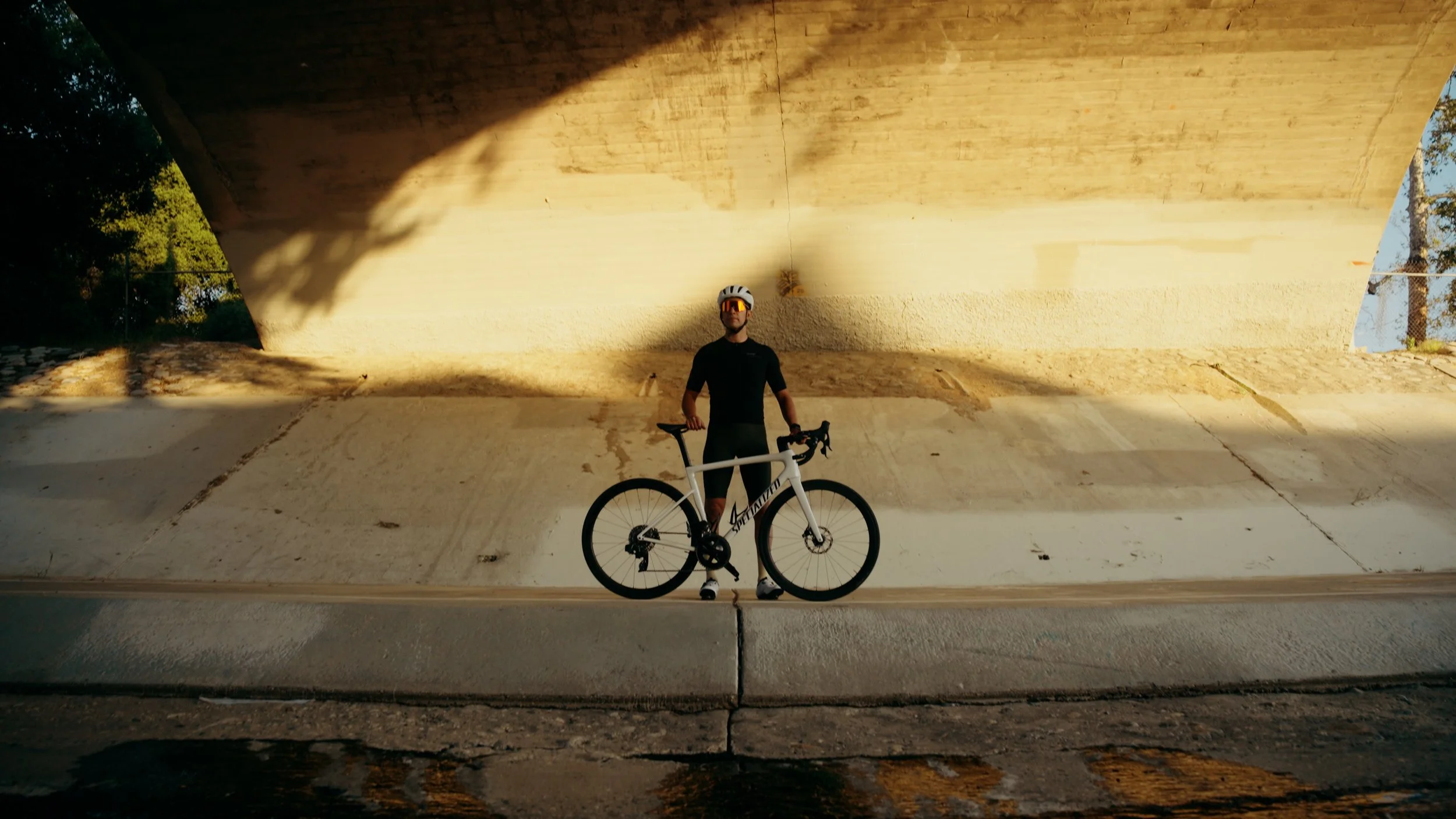 Person standing with a bicycle under a bridge at sunset, wearing cycling gear and sunglasses.