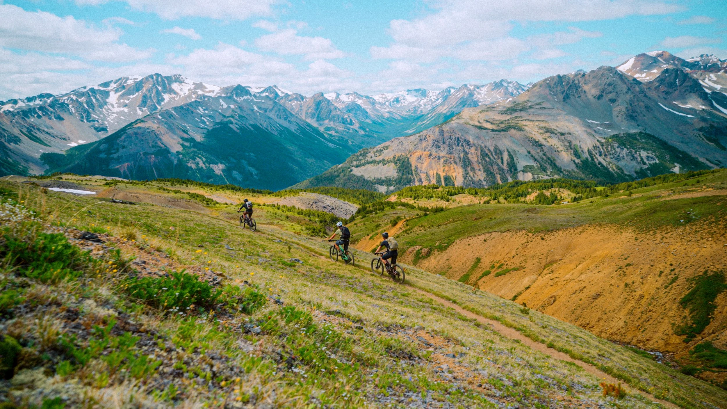 Four mountain bikers riding on a dirt trail through a green hillside with snow-capped mountains in the distance under a partly cloudy sky.