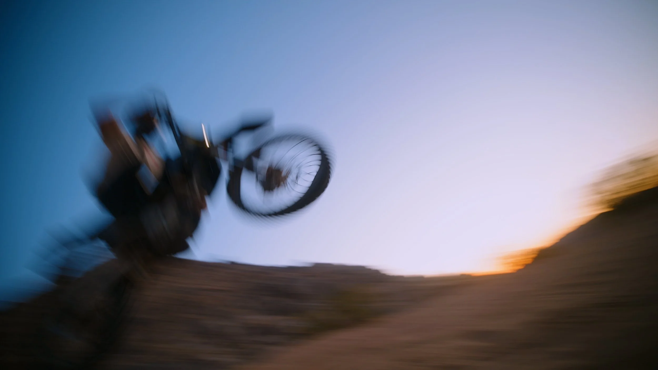 A blurred image of a person performing a stunt on a bicycle on a dirt trail during sunset.
