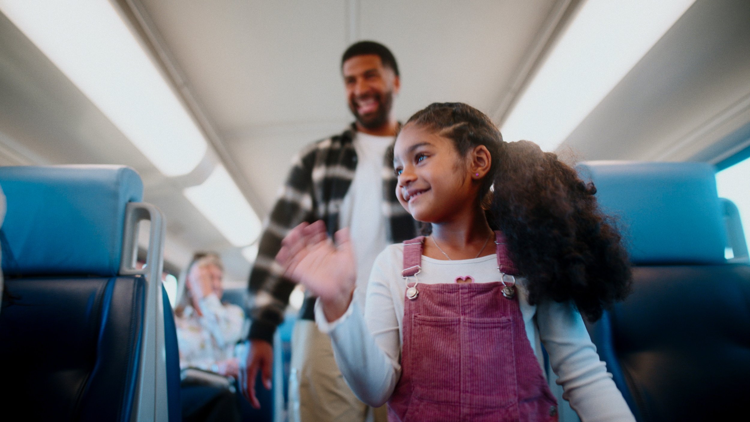 A young girl with curly hair and a pink dress waving while standing in an airplane aisle, with a smiling man in the background.