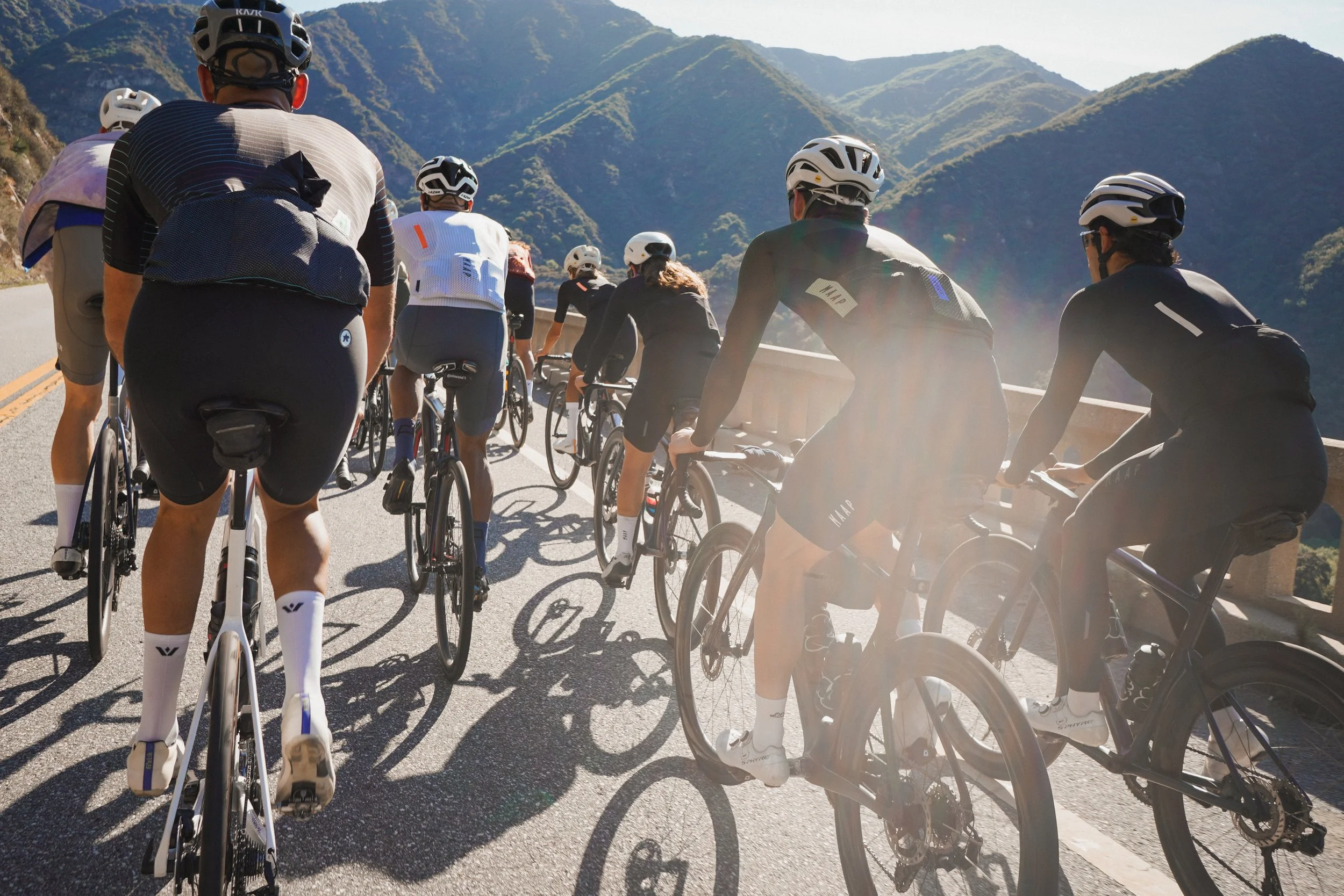 Group of cyclists riding on a mountain road with scenic mountain views in the background.