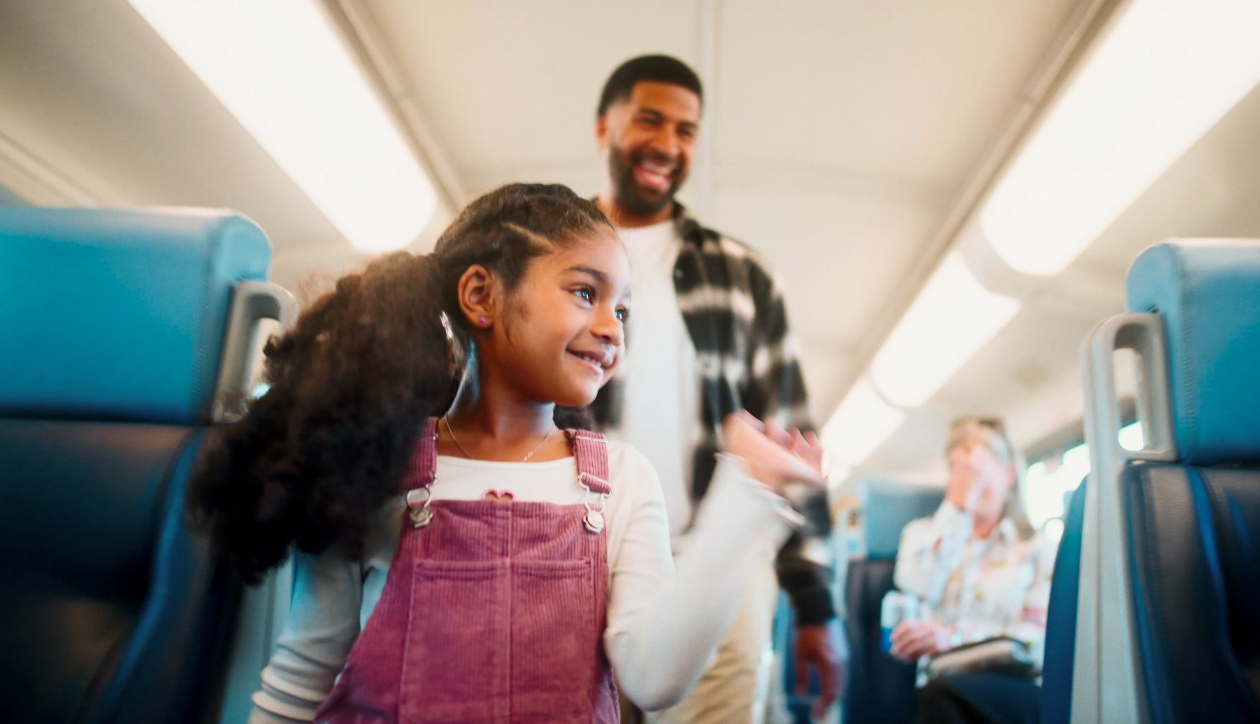 A young girl with curly hair and a pink overall smiling and waving inside an airplane cabin, with a man in a plaid shirt smiling behind her and passengers in the background.