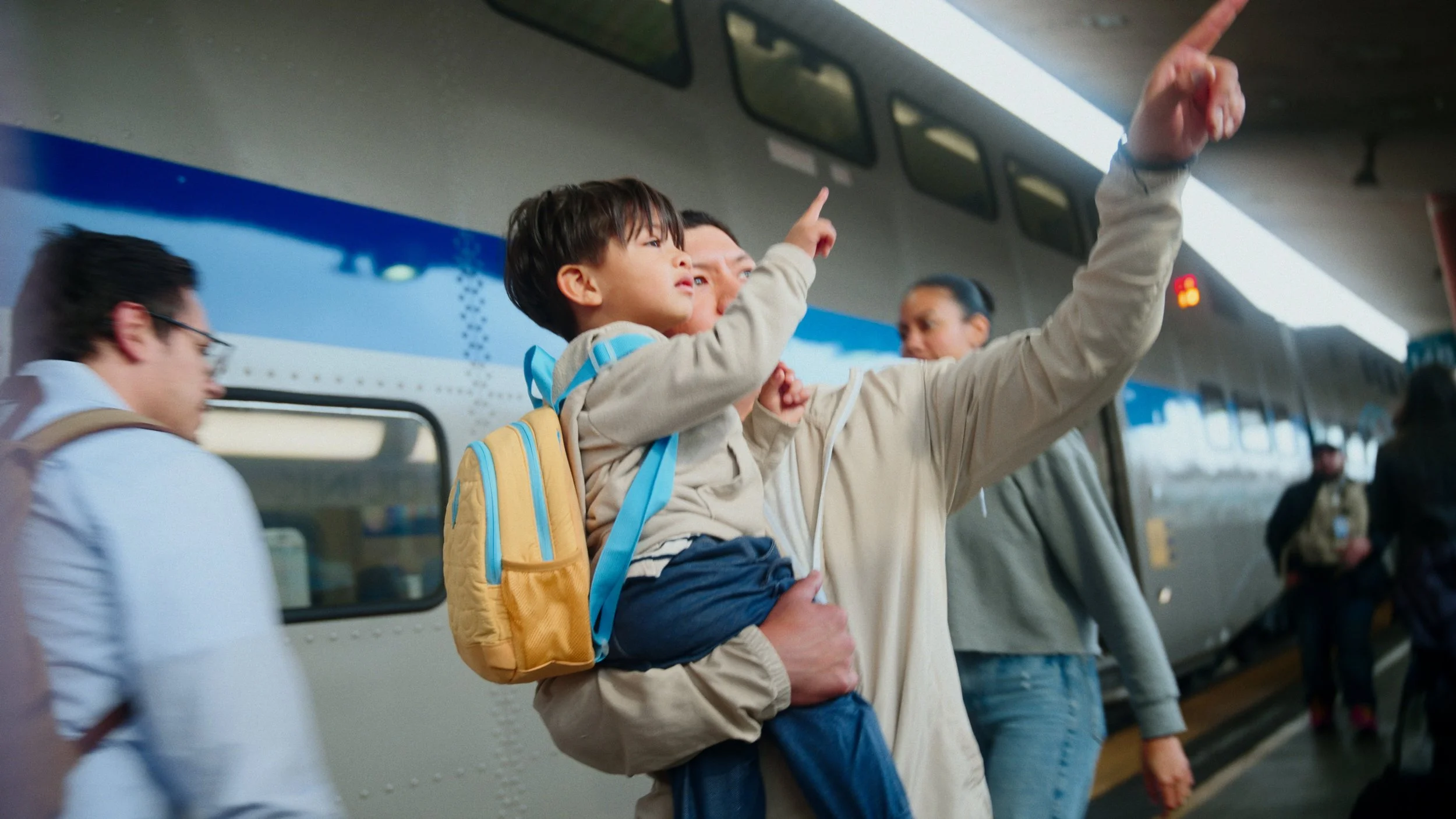 A man holding a young boy with a yellow backpack as they board a train, both pointing ahead, with other travelers in a train station.