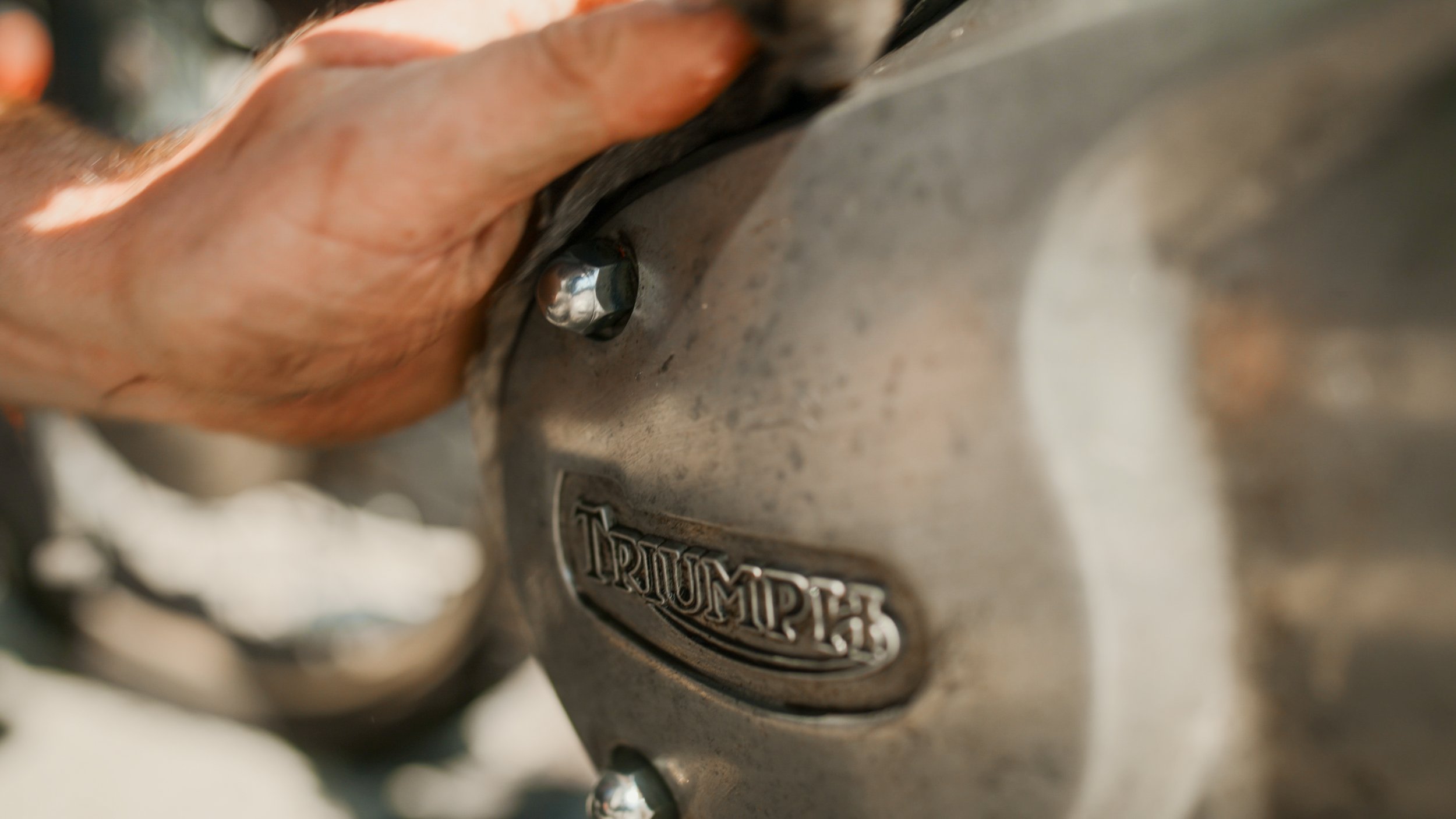 Close-up of a person's hand cleaning or working on a metallic Triumph motorcycle engine, with the Triumph logo visible.