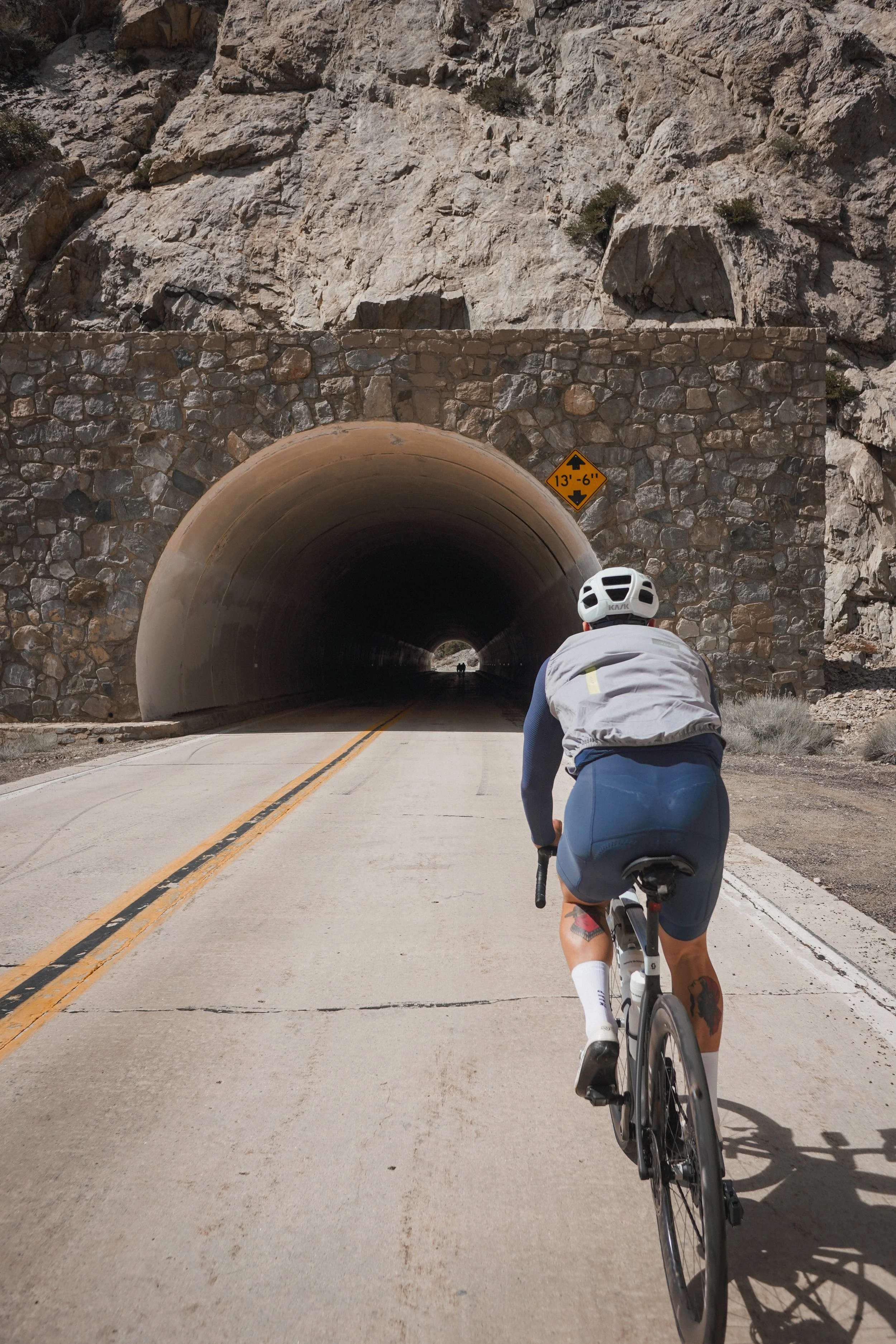 Cyclist riding toward a tunnel through a rocky mountain, with a yellow height restriction sign above the tunnel entrance.