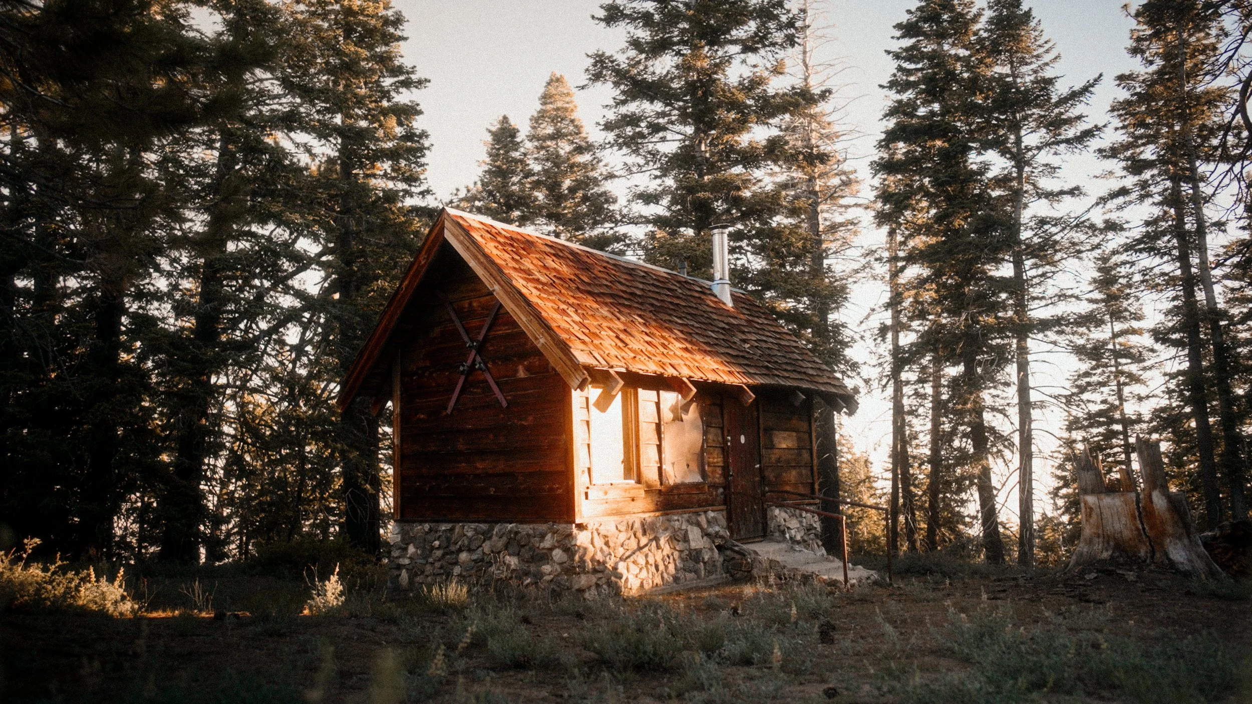 A small wooden cabin with a stone foundation situated in a forest with tall pine trees during sunset.