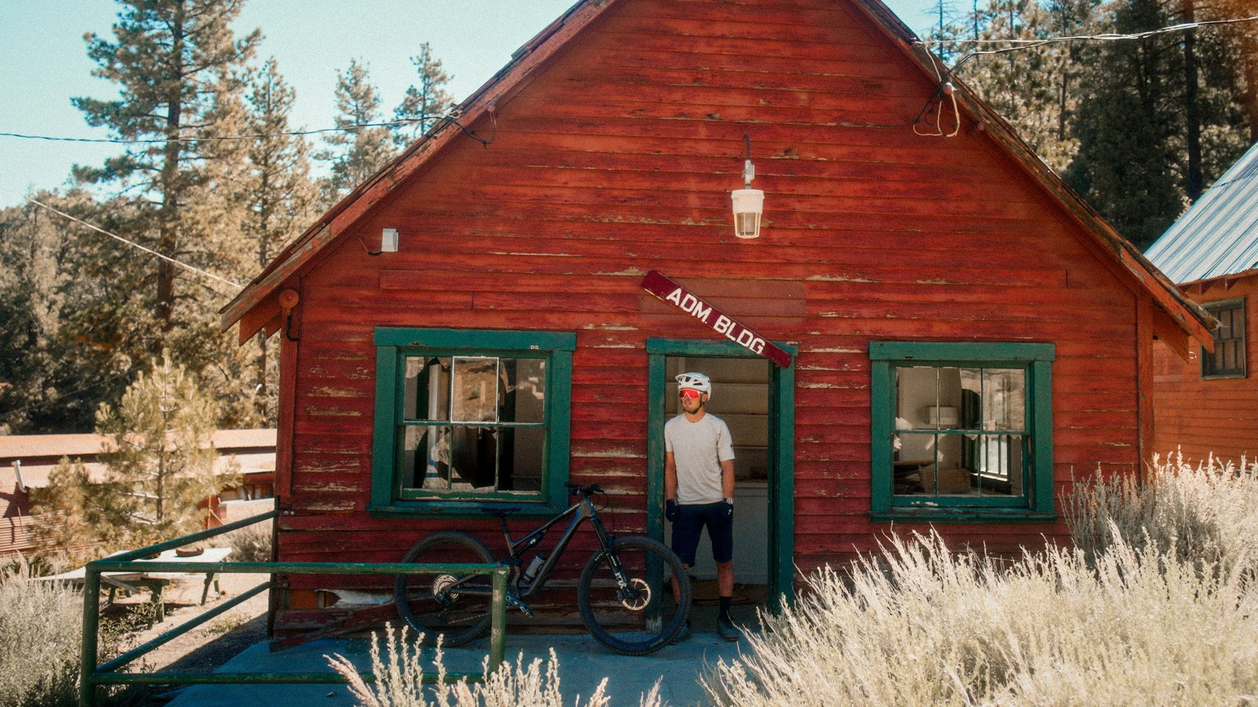 A man wearing a white helmet, sunglasses, and cycling gear stands in the doorway of a red wooden house with green window frames. A black mountain bike leans against a green railing in front of the house. The house has an 