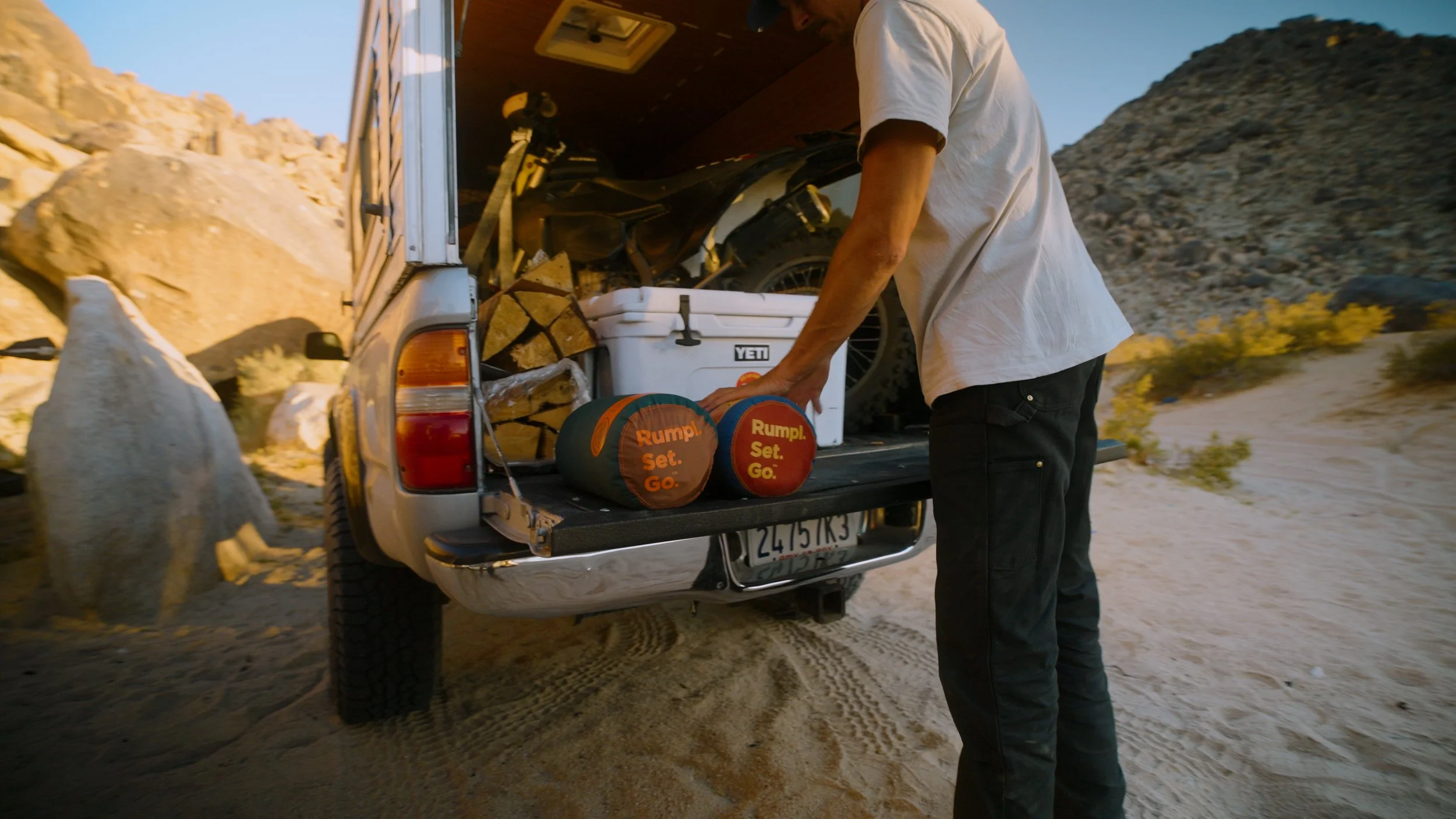 A person loading outdoor gear, including two footballs labeled 'Rumpl Set Go,' into the back of a truck in a desert area with large rocks and sand dunes.
