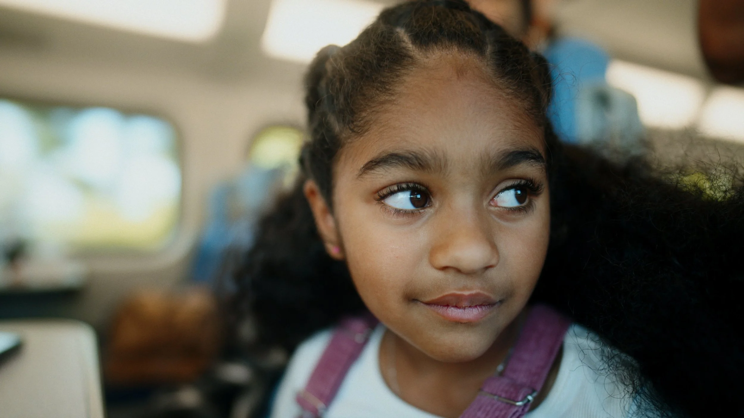 Close-up of a young girl with dark curly hair and brown eyes, wearing a white shirt and pink suspenders, inside a vehicle.
