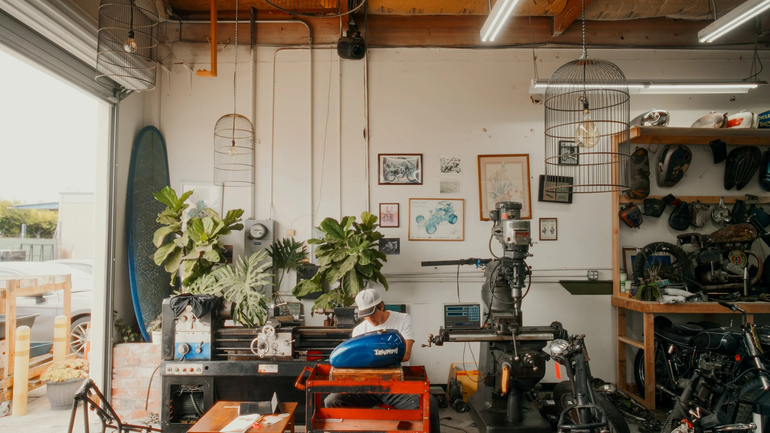 Interior of a motorcycle workshop with a person working on a blue Triumph motorcycle fuel tank. The workshop has plants, framed pictures on the wall, a large machine, a motorcycle, and shelves with helmets and motorcycle parts.