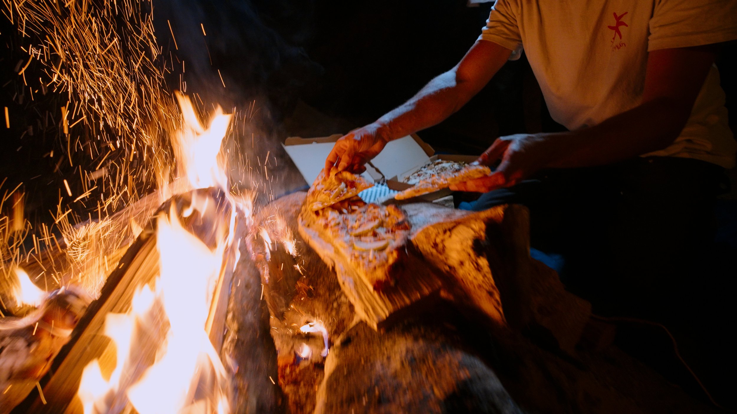 Person placing pizza slices onto a fire in an outdoor setting at night.