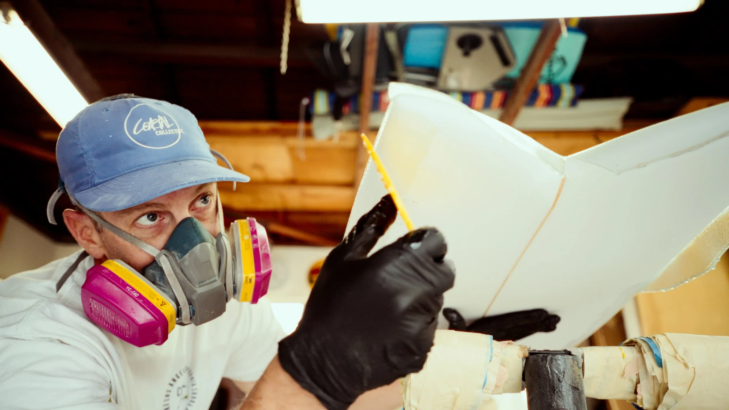A man wearing a blue cap, a respirator mask, and black gloves is painting a large white object with a yellow brush.