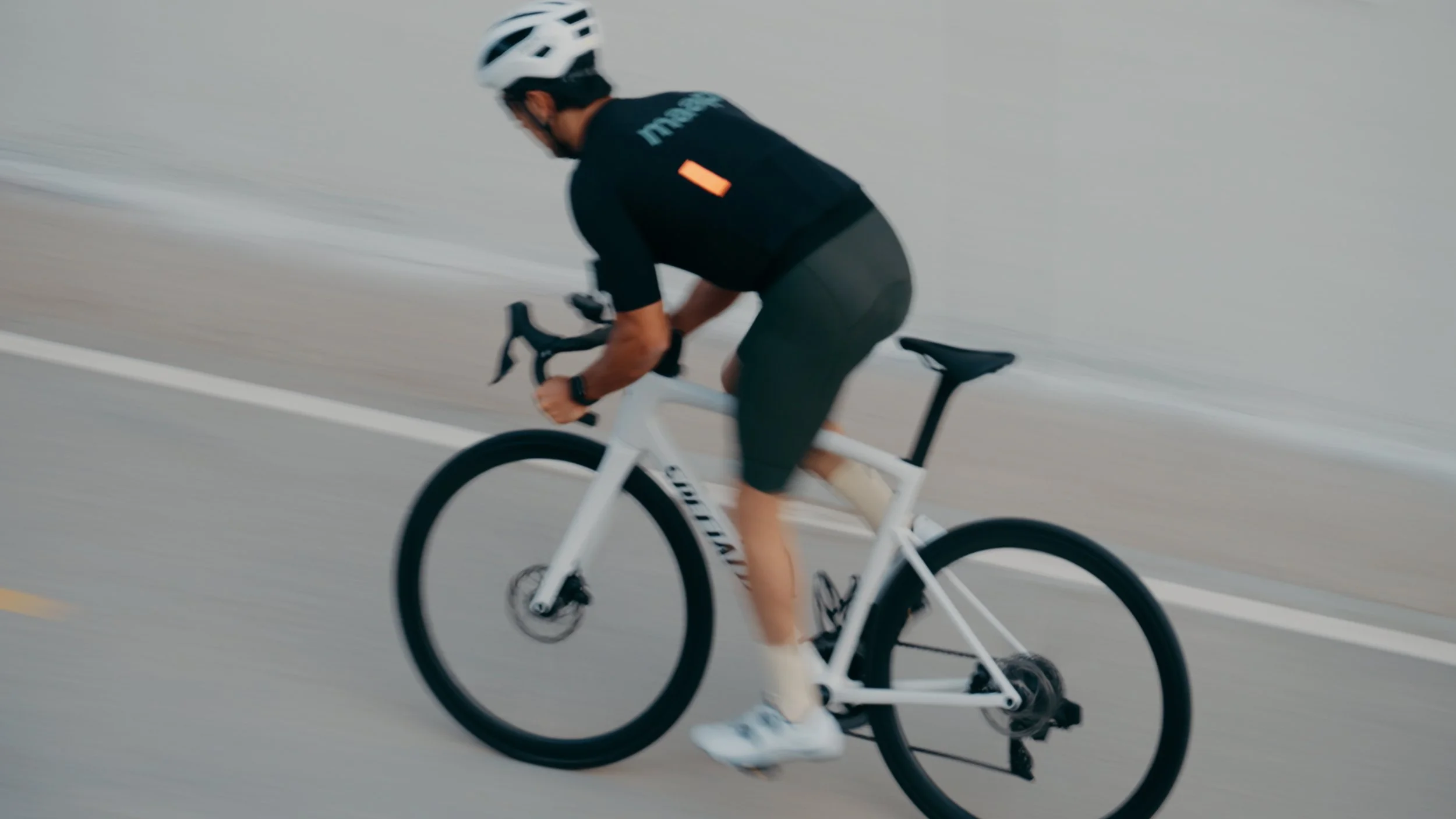 A man riding a white road bike on the street, wearing a helmet, black shirt, and gray shorts.
