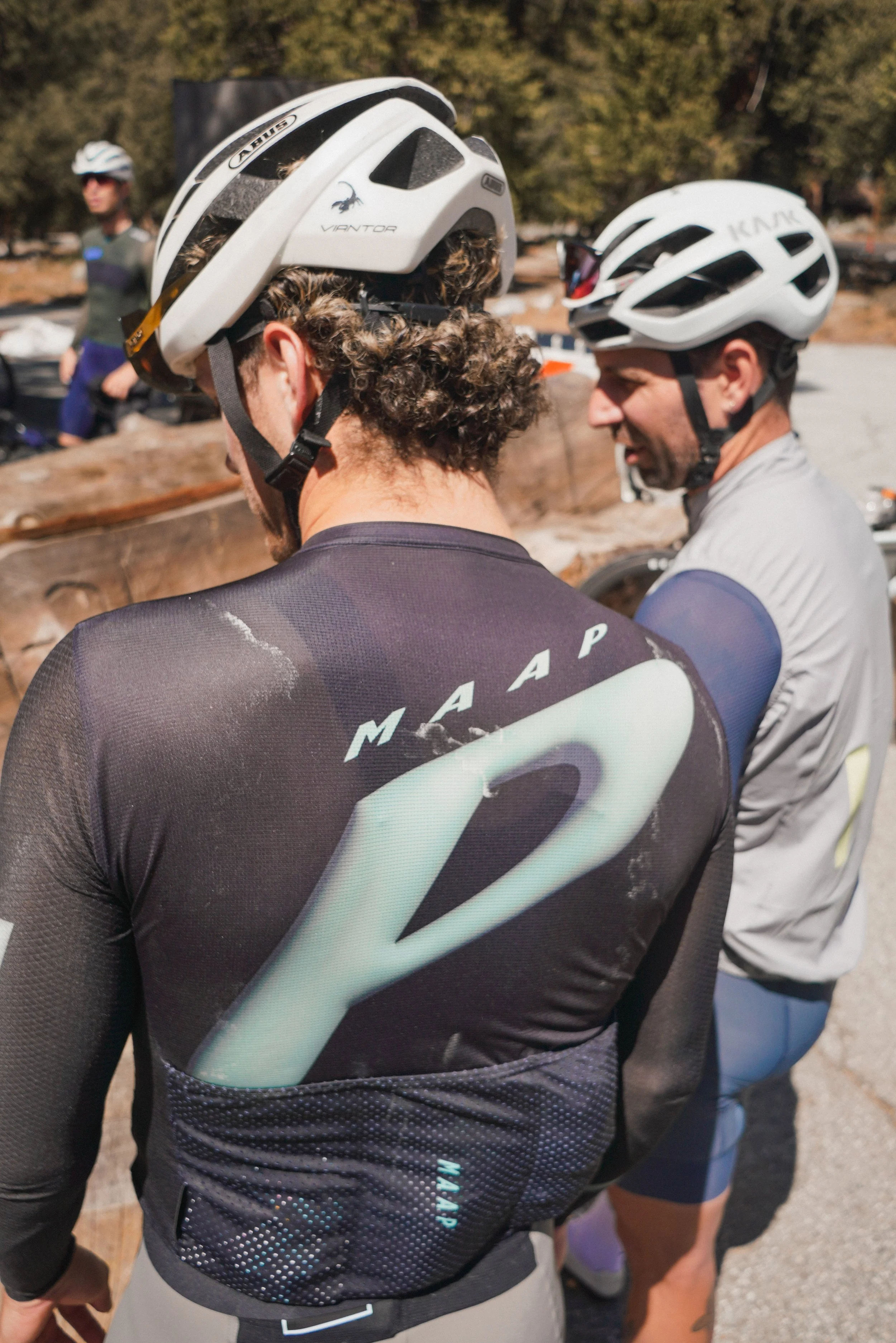 Two cyclists in helmets and athletic clothing standing outdoors, engaging in conversation, with a third person in the background.