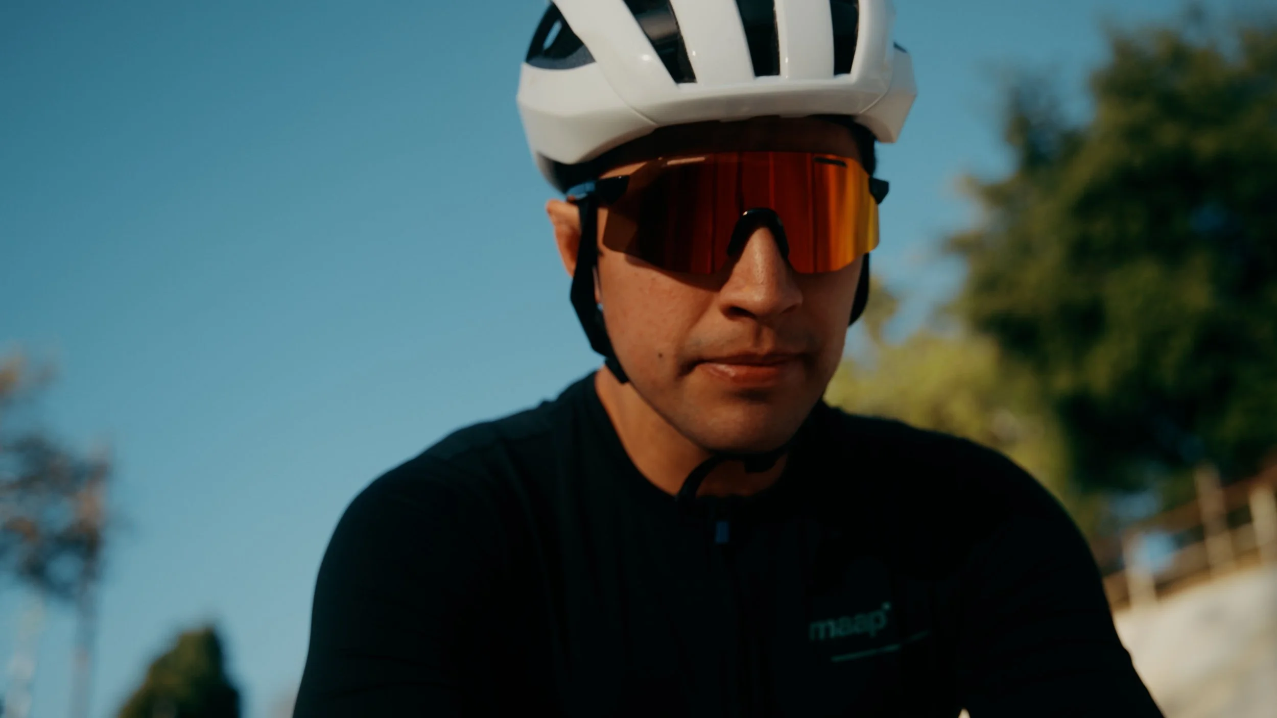 Close-up of a cyclist wearing a white helmet, orange-tinted sports sunglasses, and a black cycling shirt outdoors during daytime.