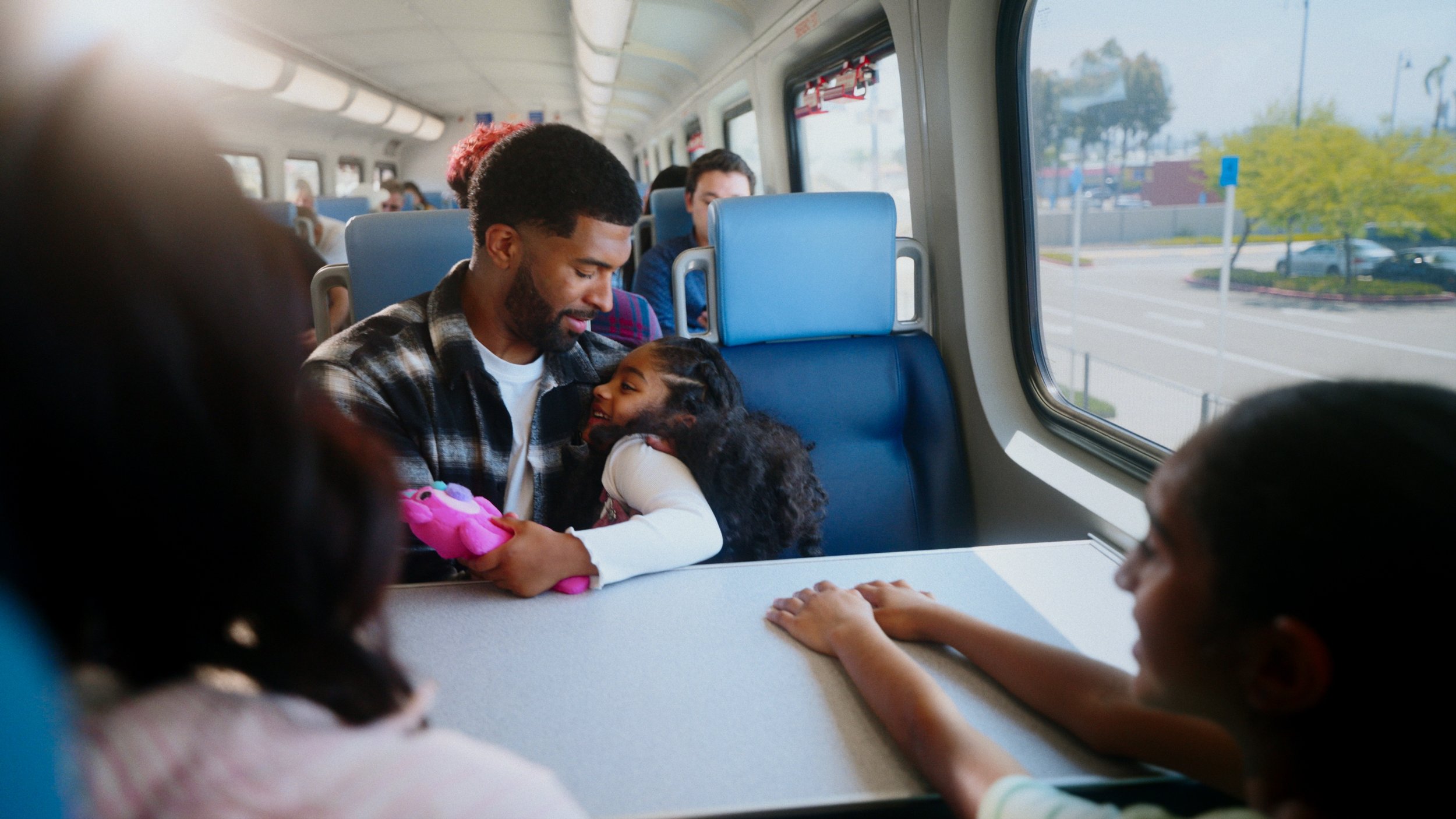 A man and a young girl with curly hair sharing a tender moment while riding on a train, surrounded by other passengers.