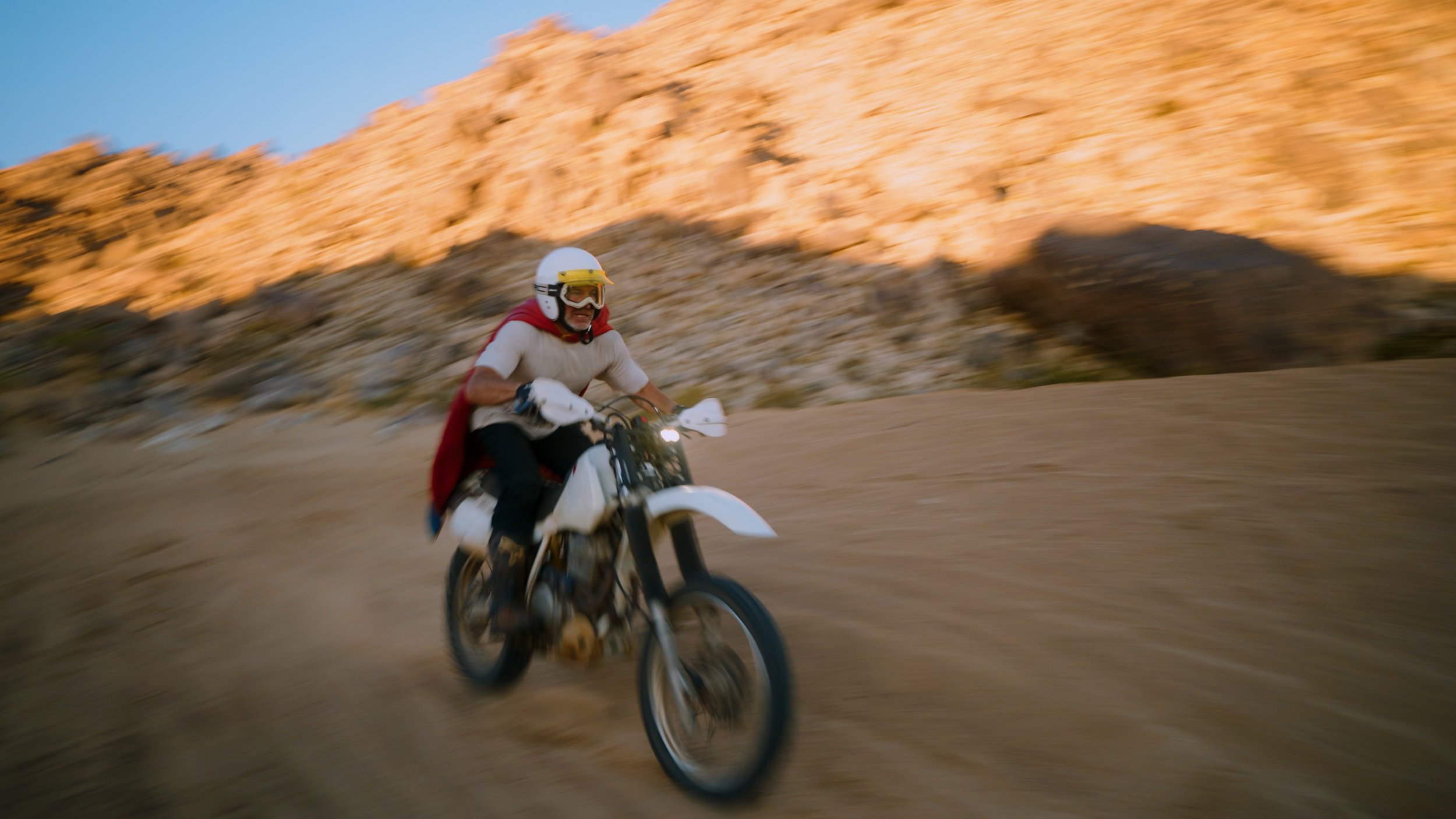 Person riding a dirt bike on a trail with desert landscape and rocky hills in the background, during sunset.