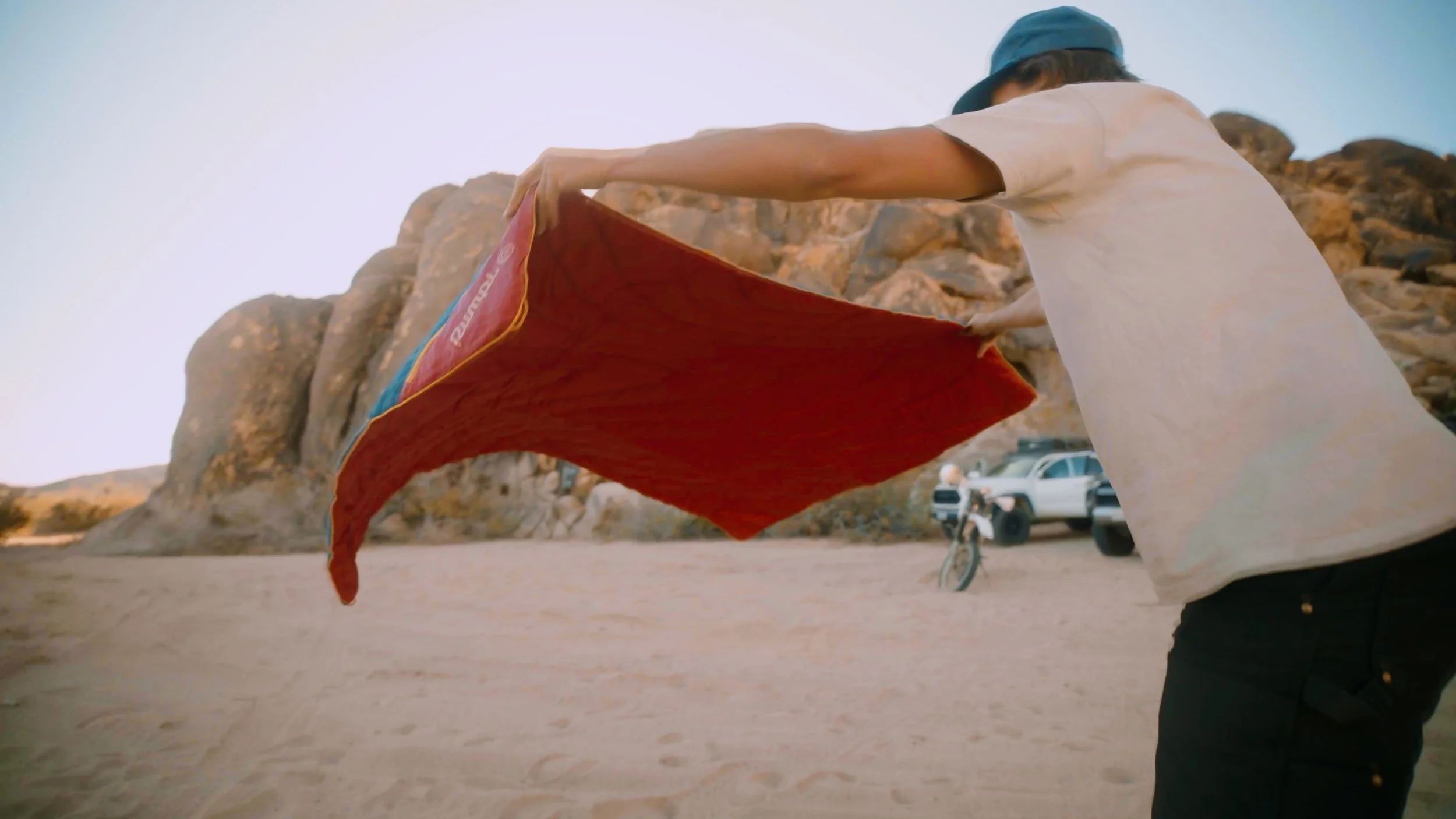 Person wearing a blue hat and beige t-shirt holding a red flag or cloth on a sandy surface with rocks and parked cars in the background.