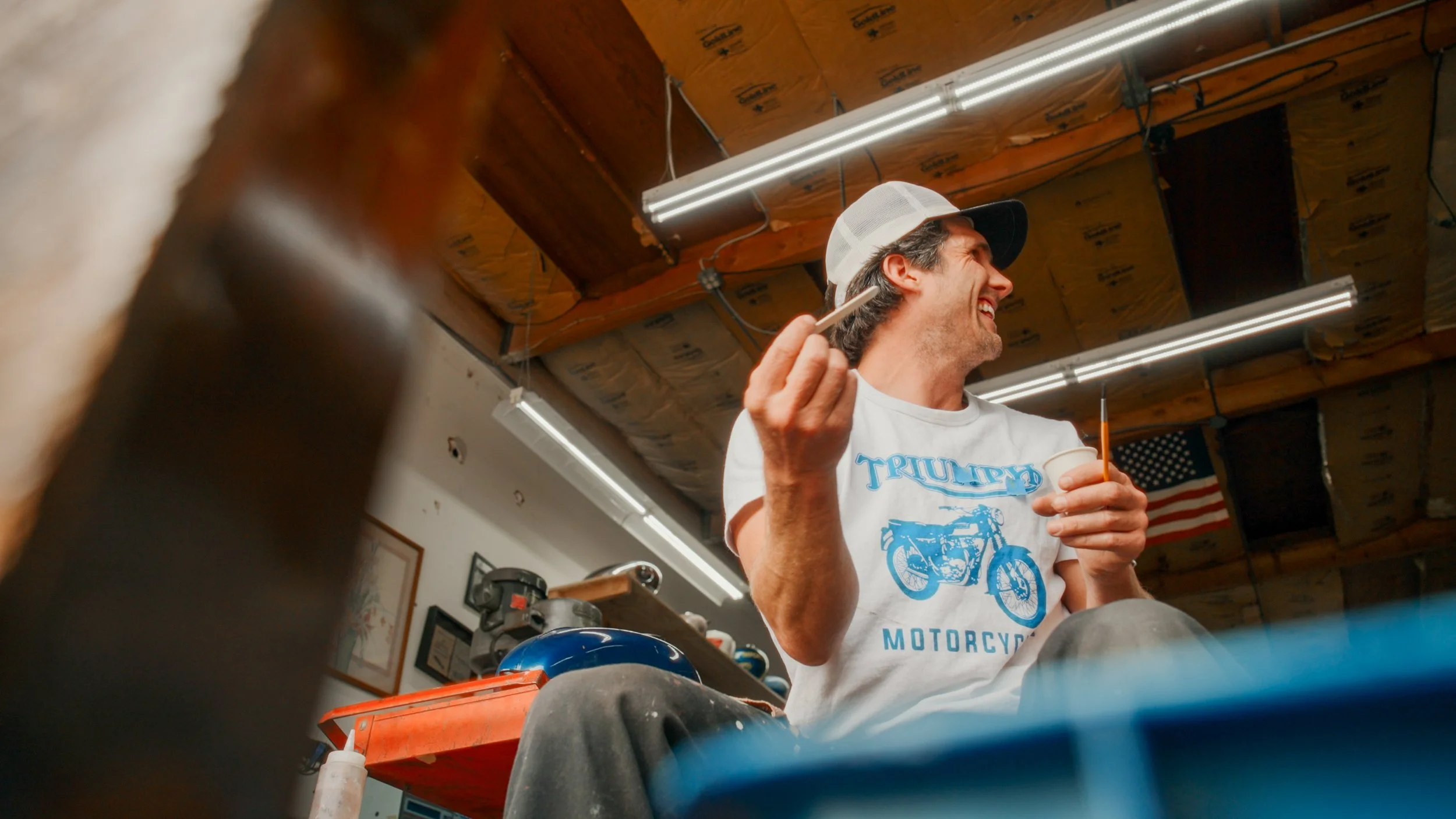 A man wearing a white T-shirt with a blue motorcycle graphic and text, a white and black baseball cap, smiling and holding a small cup and a paintbrush inside a workshop with wooden ceiling beams and fluorescent lights.
